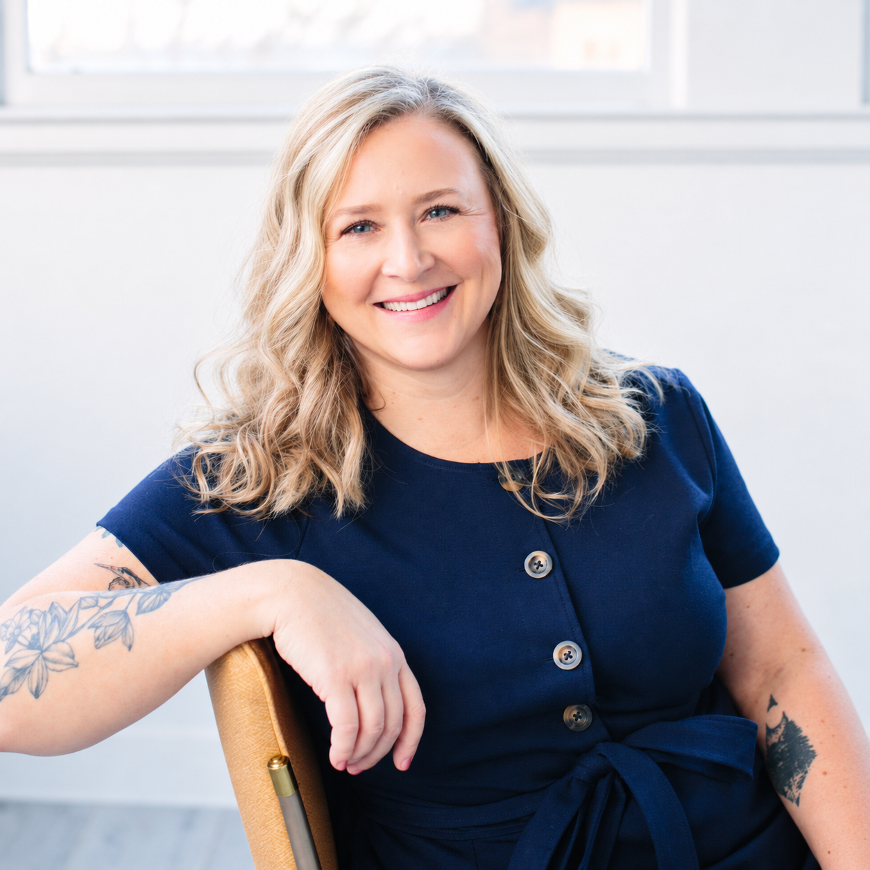 Emily Whitish, therapist and artist, smiling and wearing a navy blue dress with buttons and a drawstring waist, sitting in a light-filled room.