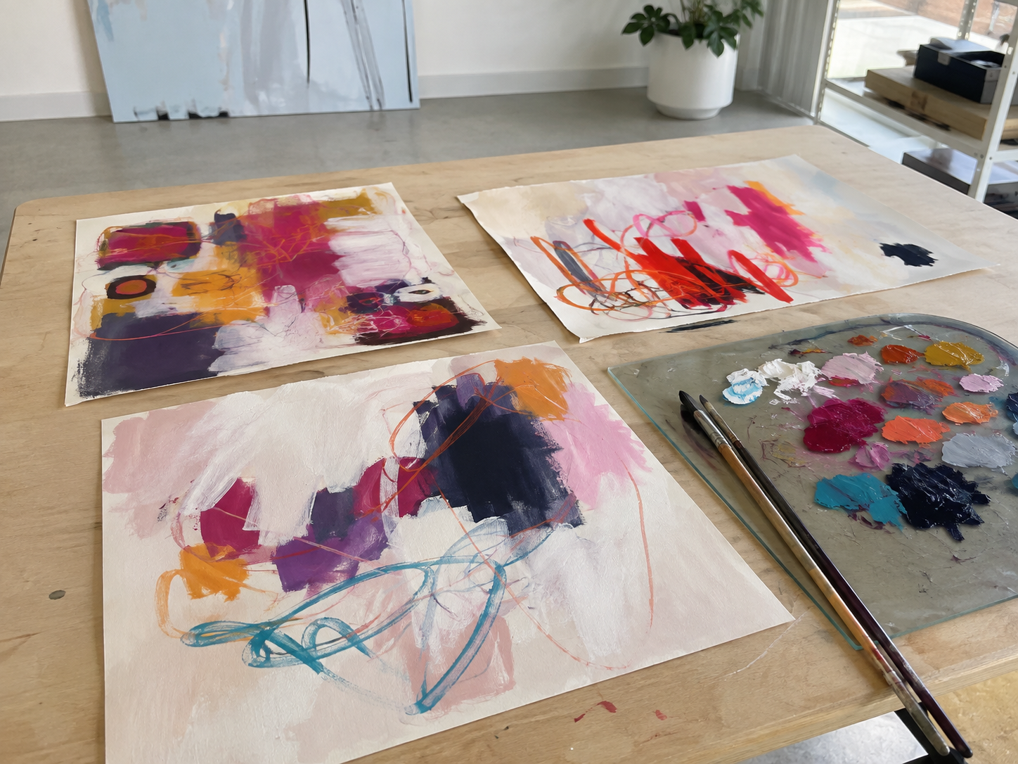 Three abstract acrylic paintings on paper spread out on a wooden studio table, with a palette of various paint colors and two brushes nearby.