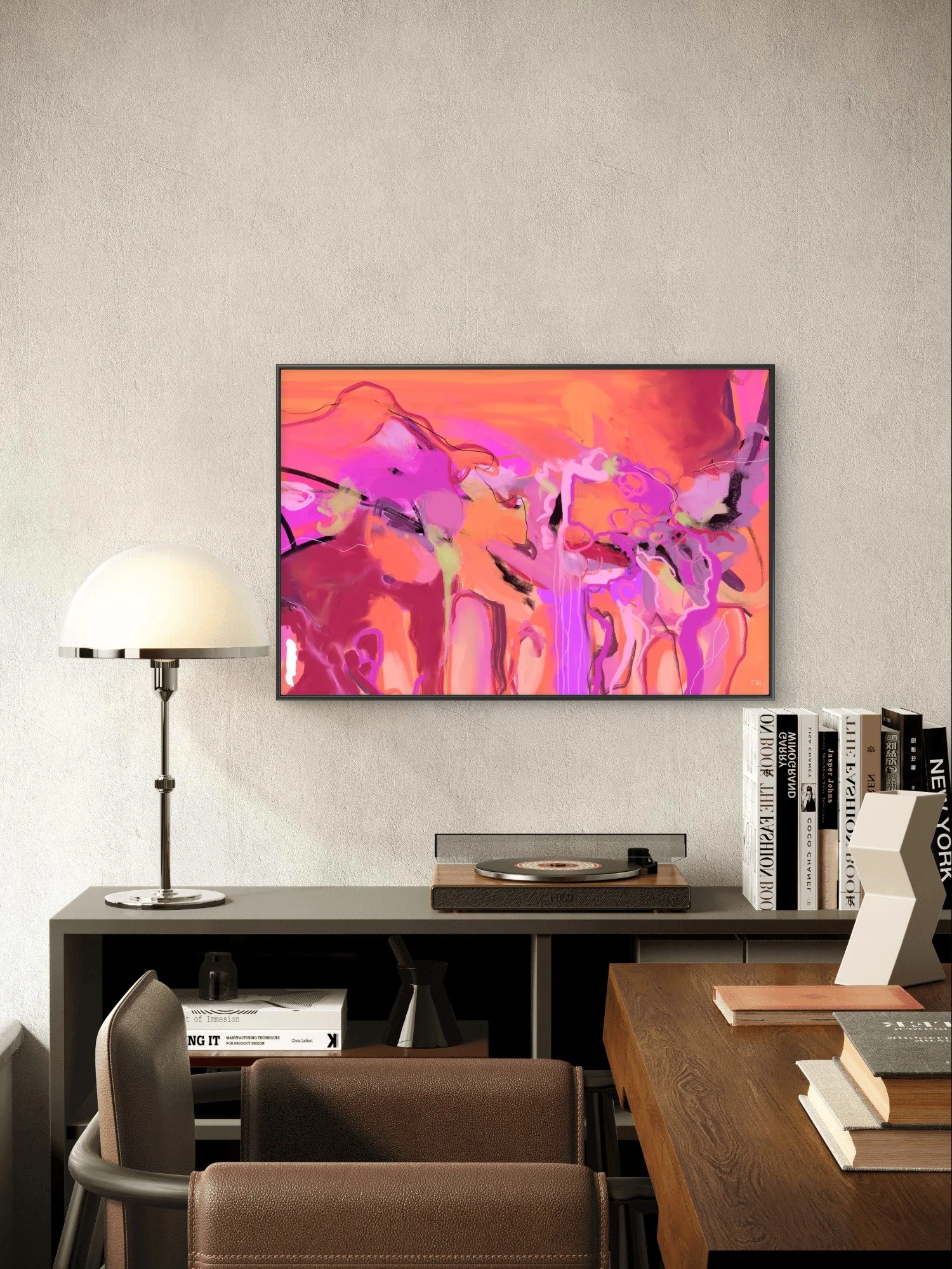 'At Home in Your Skin,' a neon pink abstract art print by Emily Whitish hangs framed in a mid-century modern living room a white table lamp, a bookshelf with art and design books, a vintage record player, and a wooden table with books and notebooks.