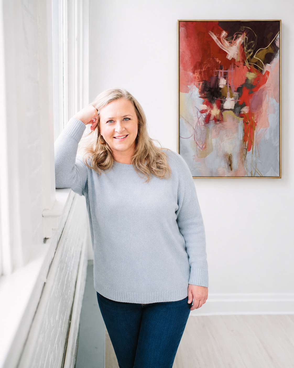 Bainbridge Island artist, Emily Whitish, standing in a studio space wearing a light sweater and blue jeans, smiling. Her abstract colorful painting,  'Still Falling' is hanging on the wall behind her.