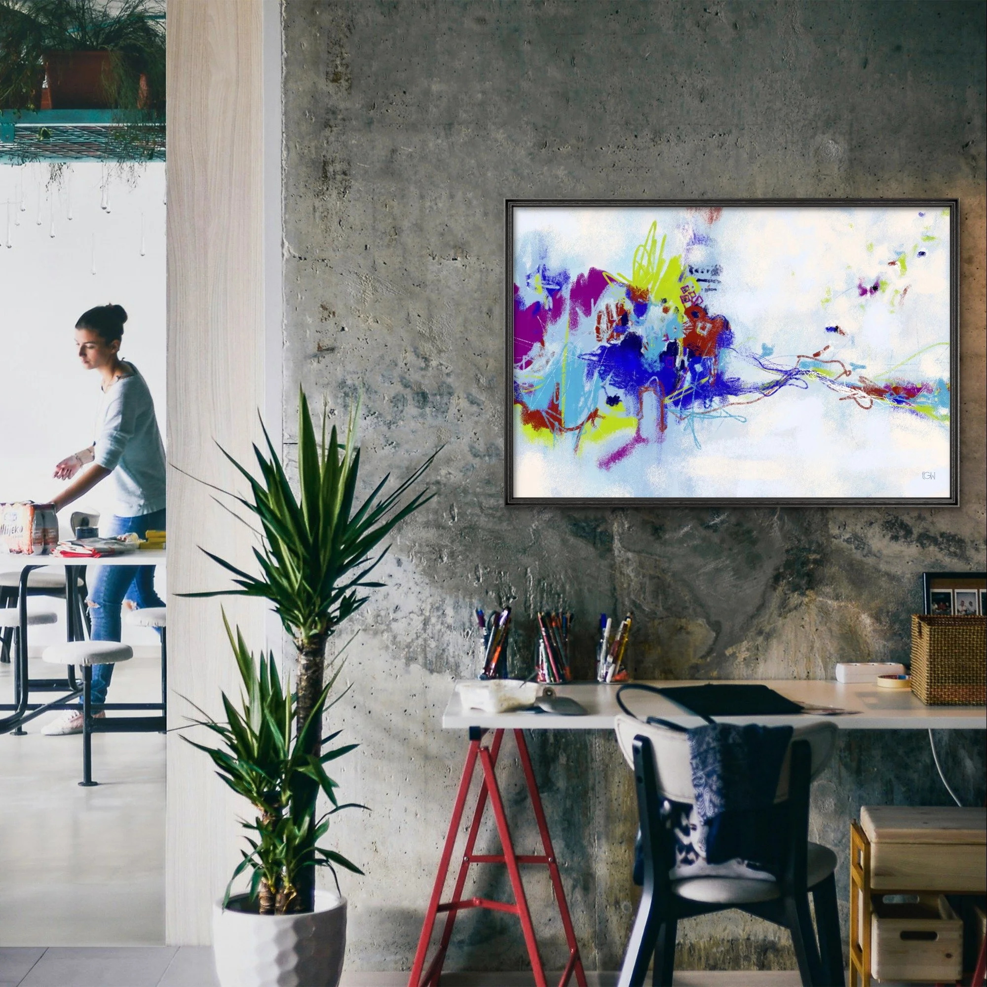 An urban tech workspace with a large potted plant in the foreground, a woman sitting at a table, and various office supplies and furniture. 'All We Have is Now,' a colorful abstract art painting by Emily Whitish hangs on the wall.