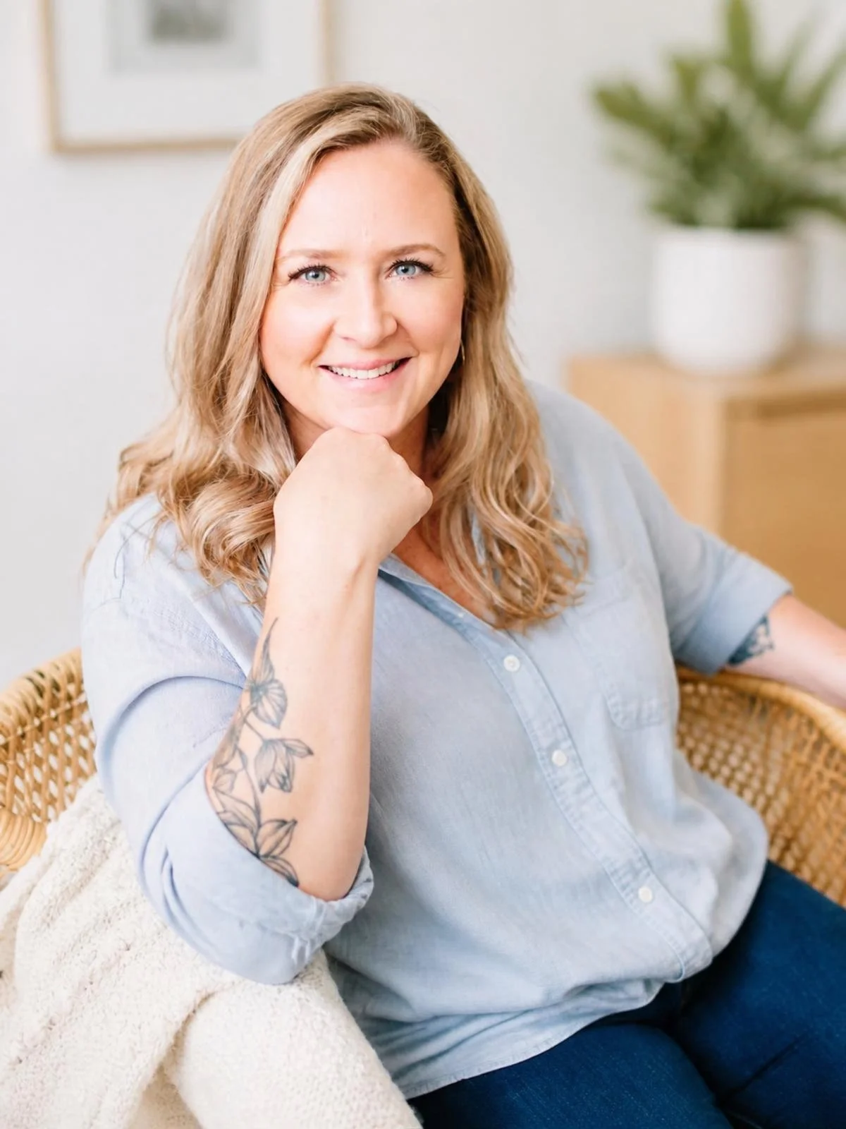 Emily Whitish, psychotherapist and abstract artist, smiling with her chin resting on her right hand, seated indoors in front of a plant and framed artwork.