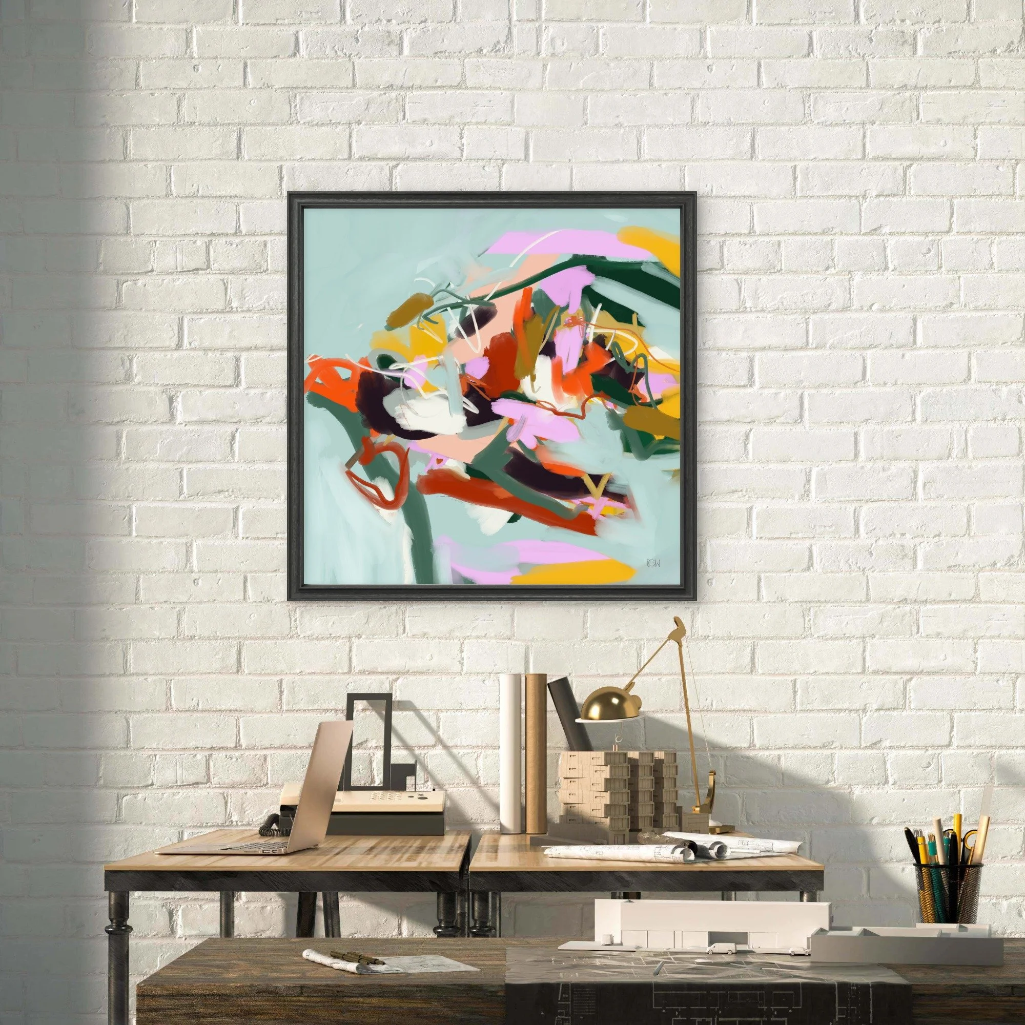 A home workspace with a desk, laptop, architectural design tools, and a framed abstract painting called 'Fly Little Bird' by Emily Whitish hangs on a white brick wall.