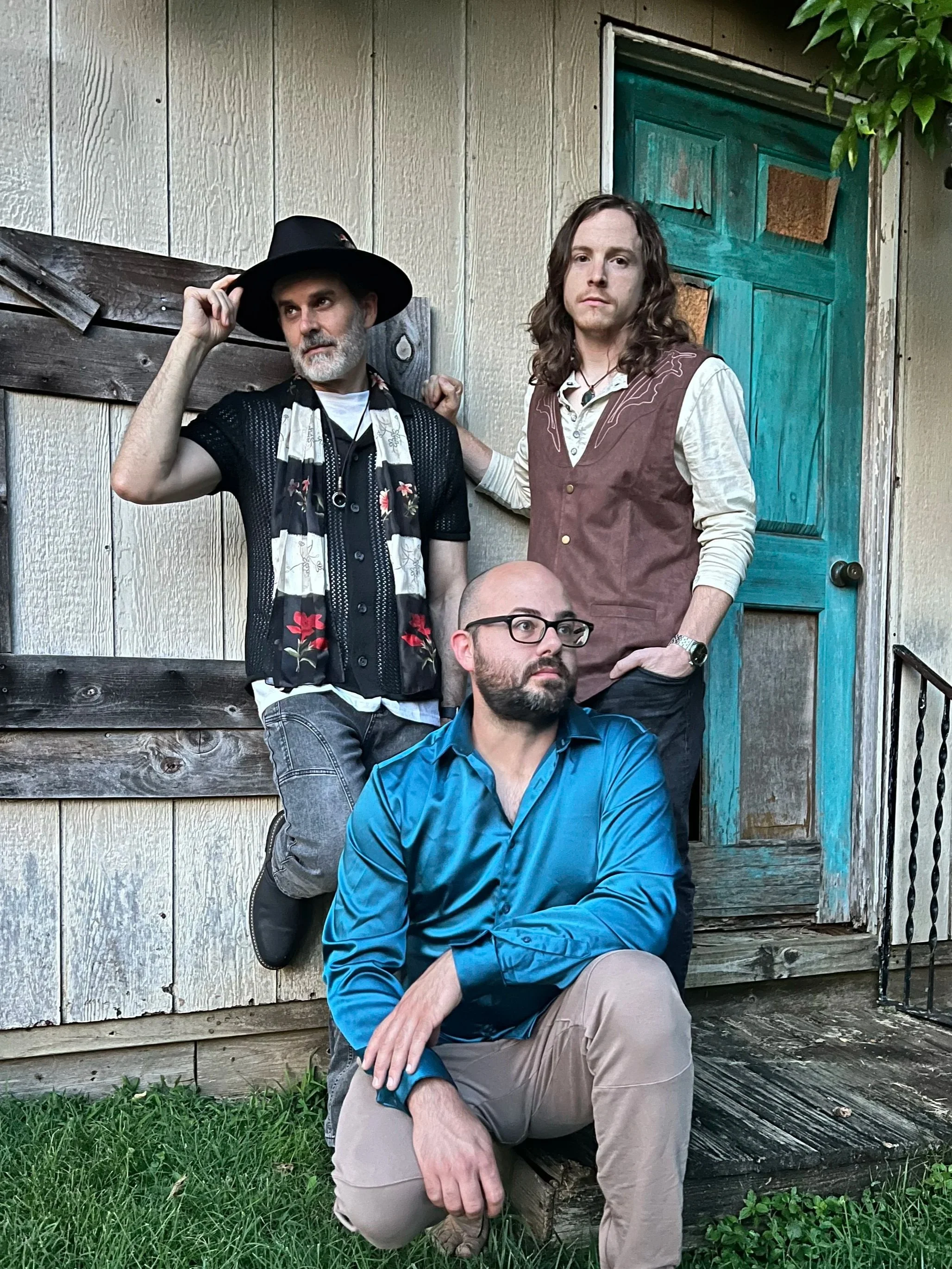 The members of the band Soundabout Revival, Hughes Taylor, Ben Alford, and Nich Gannon, standing in front of a rustic wooden building wearing vintage and casual style clothing.