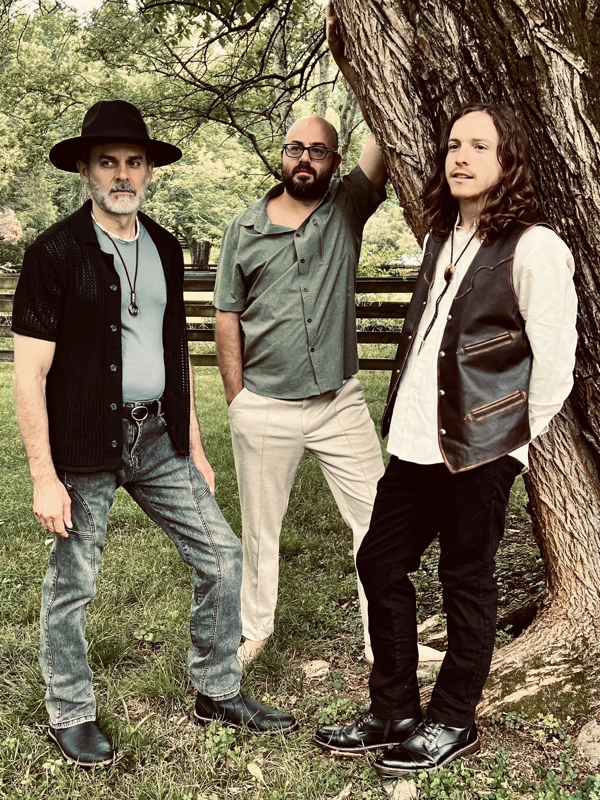 The members of the band Soundabout Revival, Hughes Taylor, Ben Alford, and Nich Gannon, standing outdoors next to a tree, dressed in casual and vintage style clothing, with green trees and grass in the background.