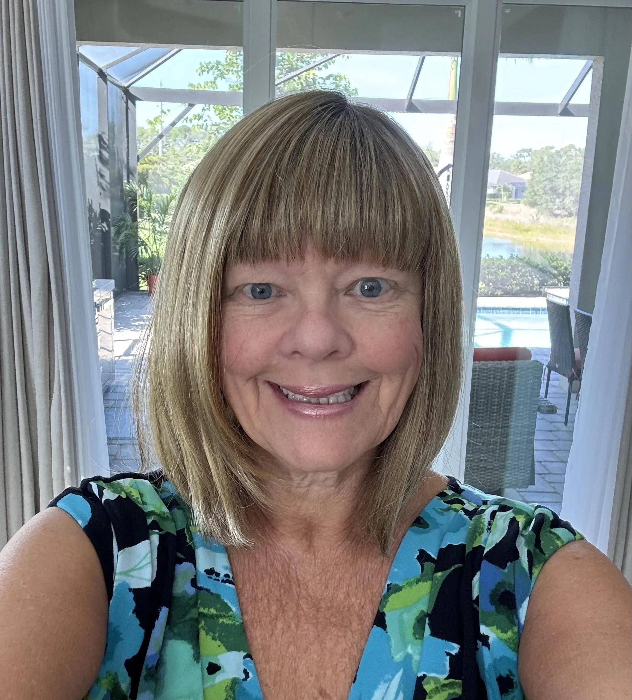 Close-up selfie of a smiling woman with shoulder-length blonde hair and blue eyes, wearing a colorful, floral-patterned top. Behind her is a patio with outdoor furniture, a pool, and a view of trees and houses through sliding glass doors.