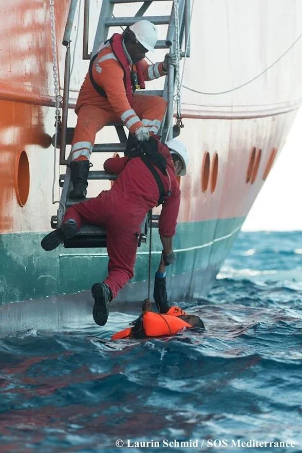 A person in a red jumpsuit is hanging onto the side of a large ship, while another person in an orange jumpsuit and helmet is on a ladder helping or rescuing them, with a small boat or flotation device in the water below.