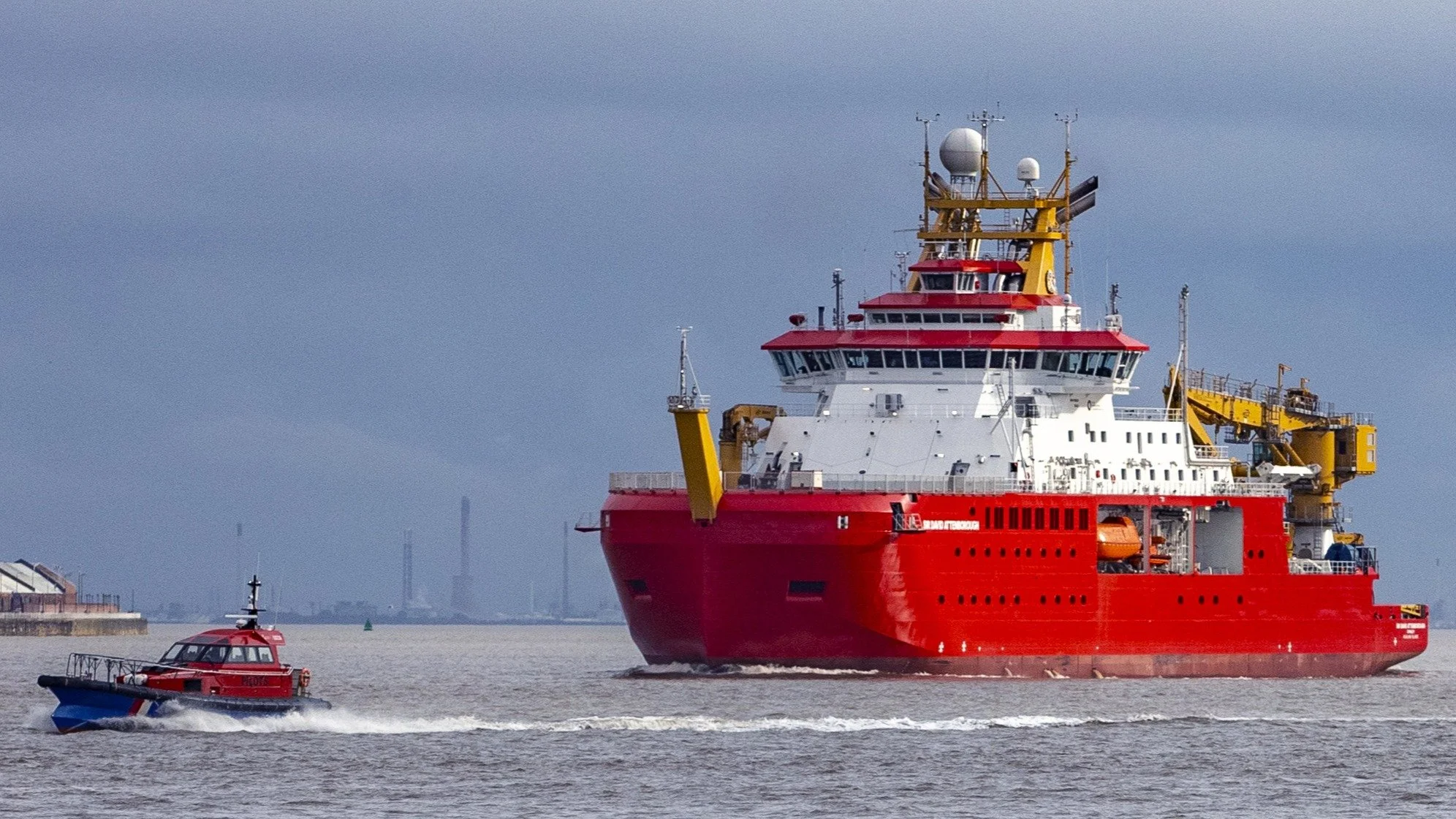 A large red ship with white and yellow structures navigates through a body of water, accompanied by a smaller red and blue boat nearby, with a city skyline in the background under a cloudy sky.