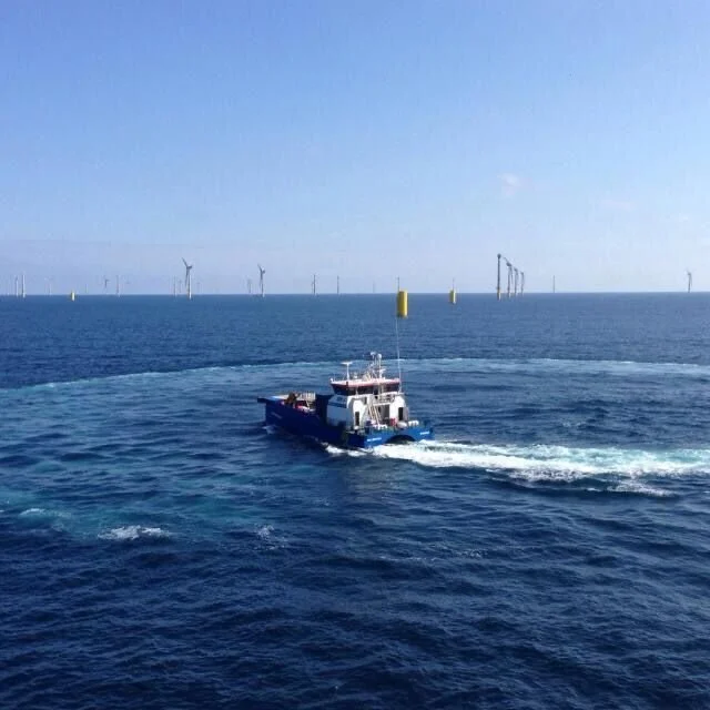 A boat moving across the open ocean with wind turbines in the background under a clear blue sky.