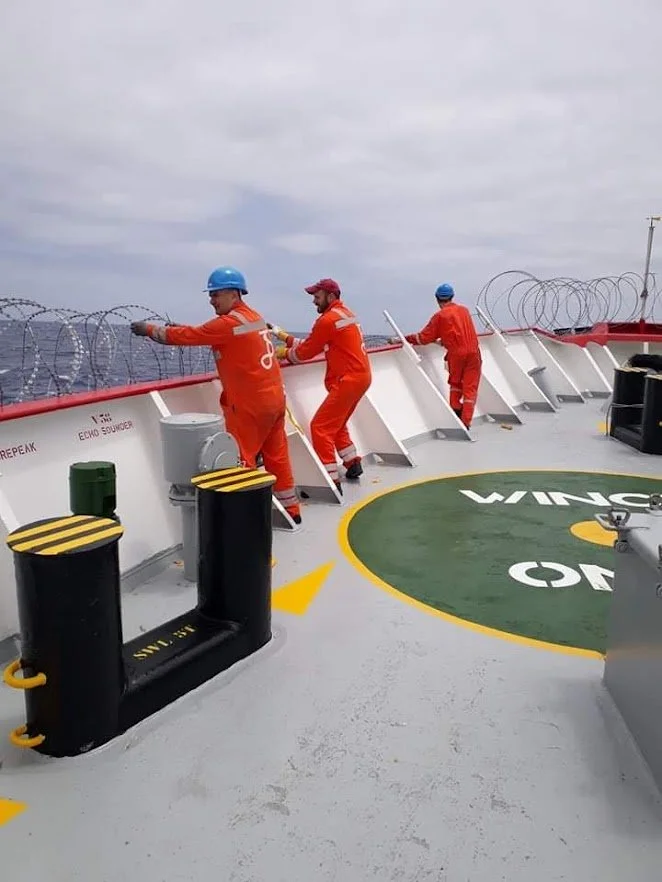 Four sailors in orange uniforms and blue helmets working on the deck of a ship, with barbed wire along the edge and overcast sky in the background.