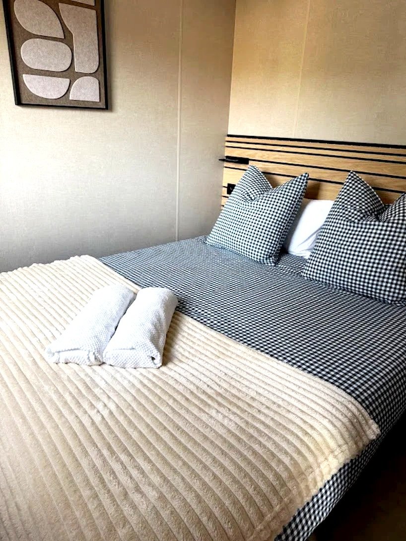Lit avec housses à carreaux en noir et blanc, oreillers assortis, et une couverture beige à rayures. Mur beige avec une œuvre d'art abstraite et un headboard en bois.