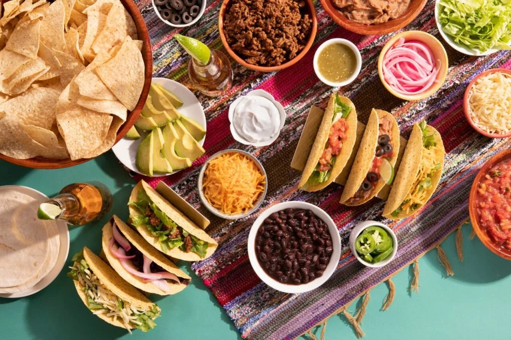 A Mexican taco feast with various tacos filled with meat, vegetables, and cheese, along with bowls of beans, shredded cheese, chopped lettuce, sliced avocados, lime wedges, chips, and condiments on a colorful woven tablecloth.