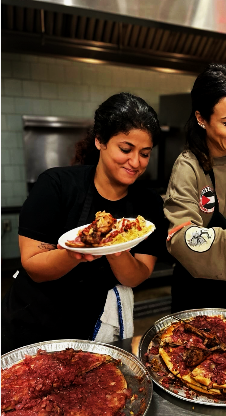 A woman holding a plate with pasta, chicken, and vegetables in a kitchen setting, with two pizzas on the counter in front of her.