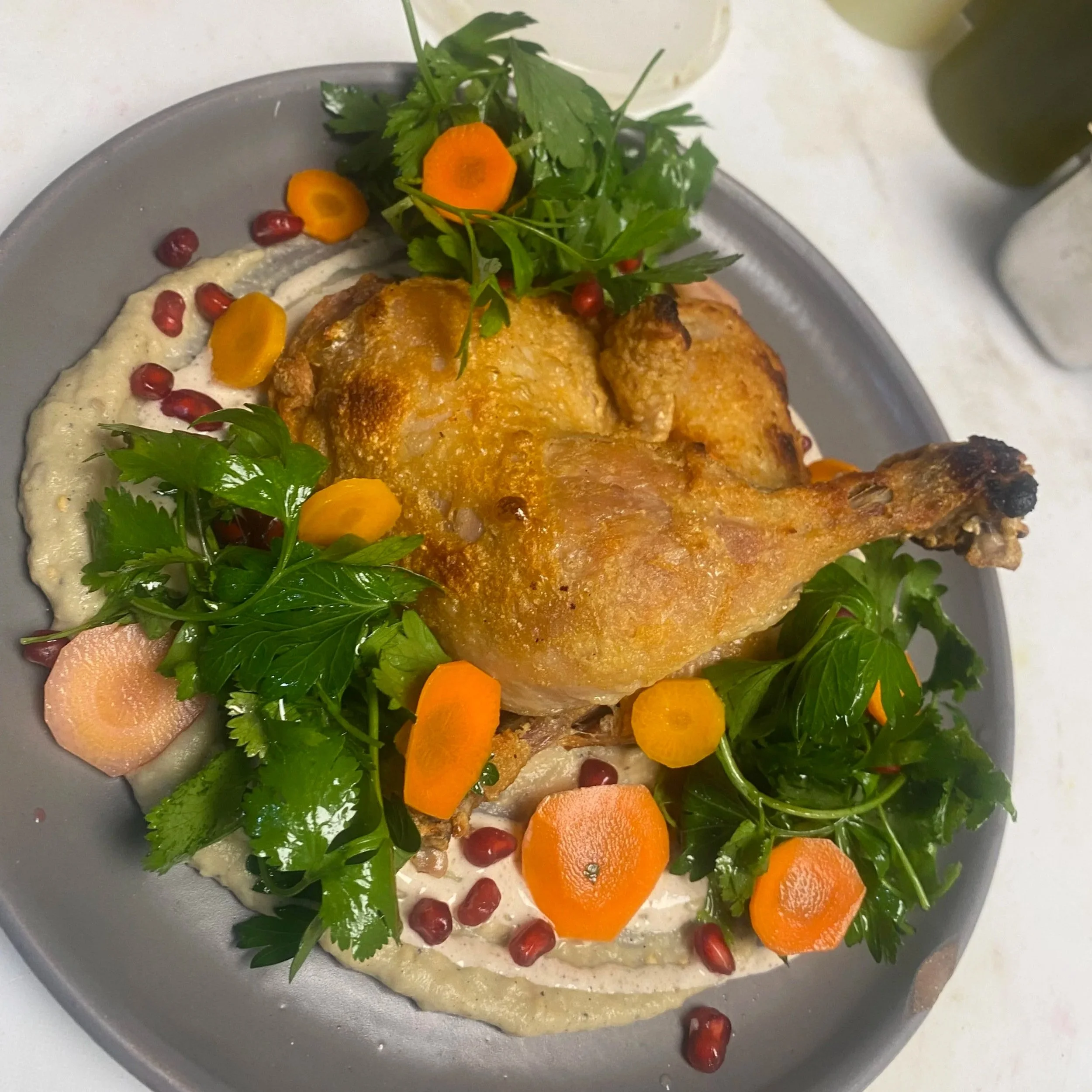 A plate of roasted chicken topped with orange slices, surrounded by pomegranate seeds, fresh parsley, and a creamy white sauce, with a side of leafy greens and a drink in the background.