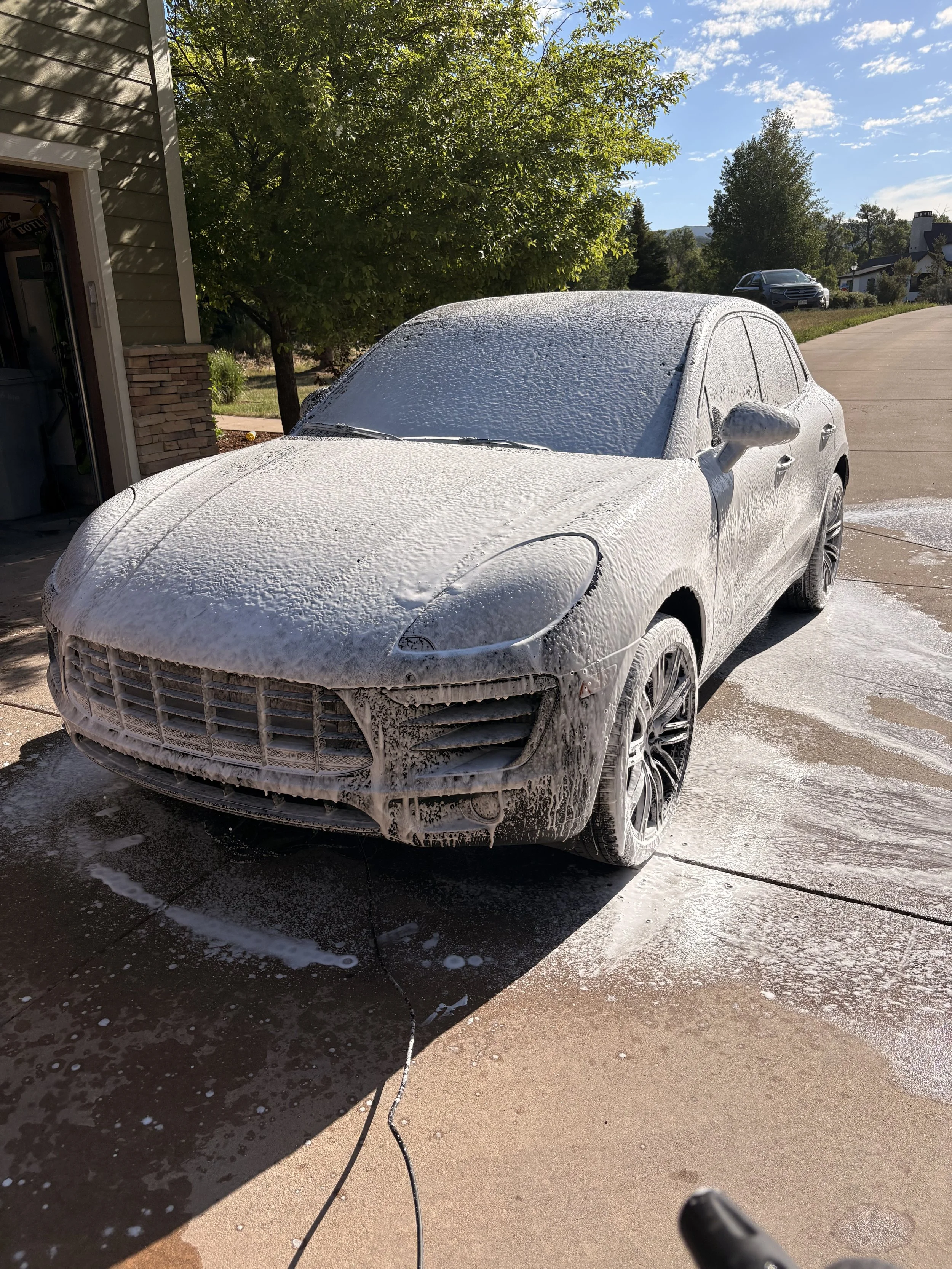 A white car covered in soap foam during a car wash in a driveway on a sunny day, with trees and houses in the background.