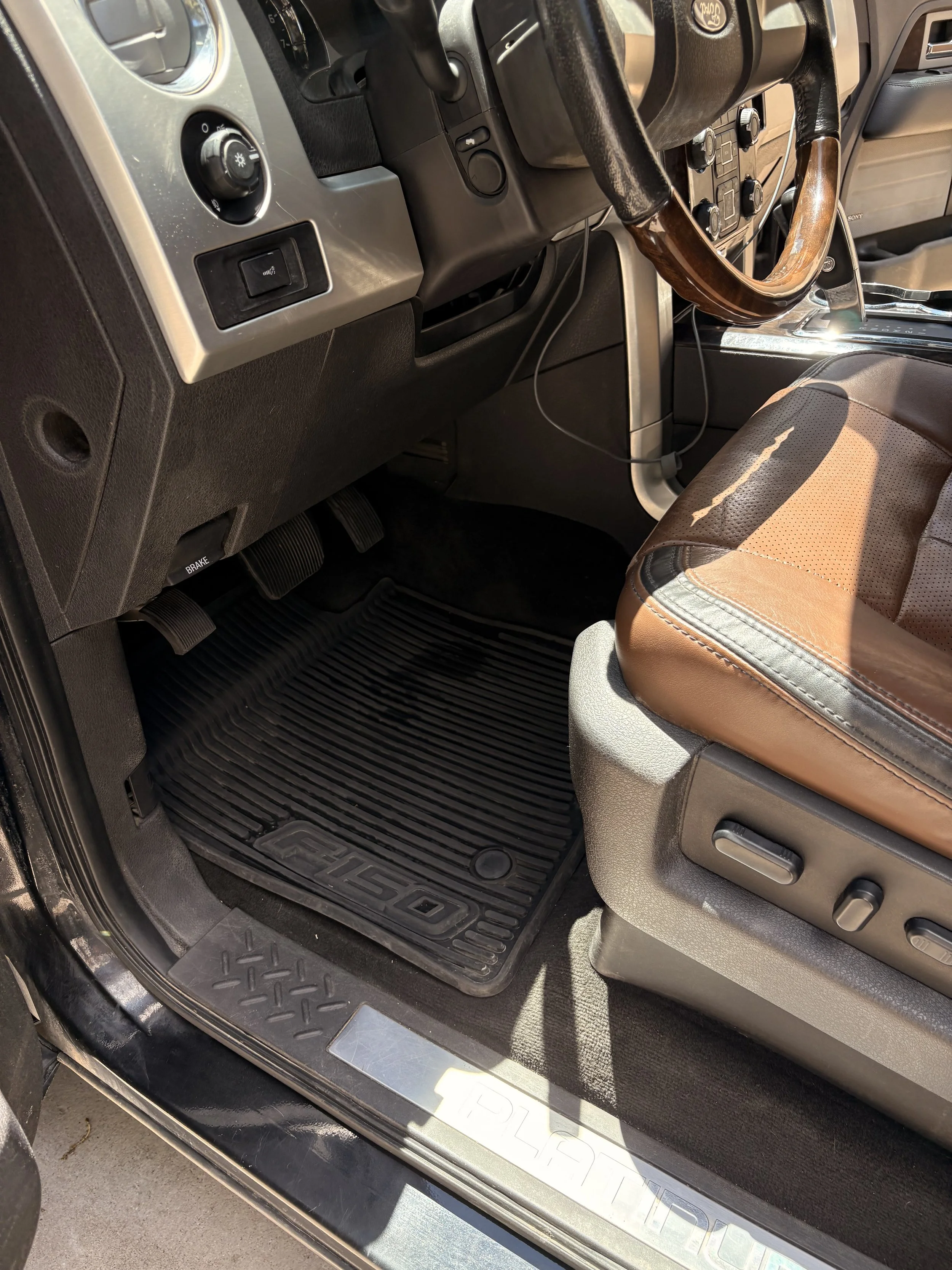 The interior of a vehicle showing the driver's side, including the steering wheel, dashboard, gas and brake pedals, and a brown leather seat. A black floor mat with a textured surface is visible on the floor.