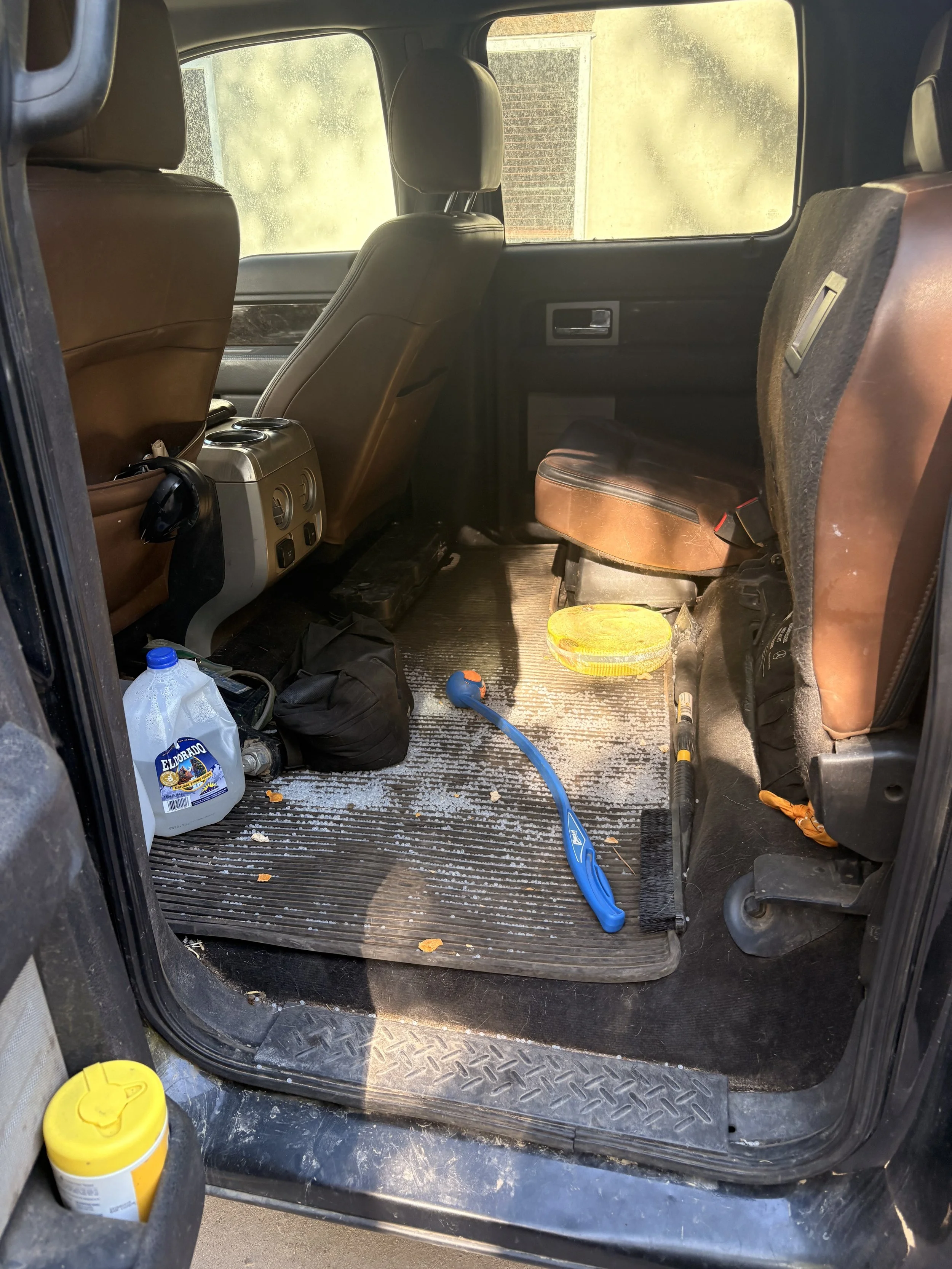 Interior of a vehicle's back seat area with a dirty floor mat, scattered trash, a yellow jar, and a blue hose.
