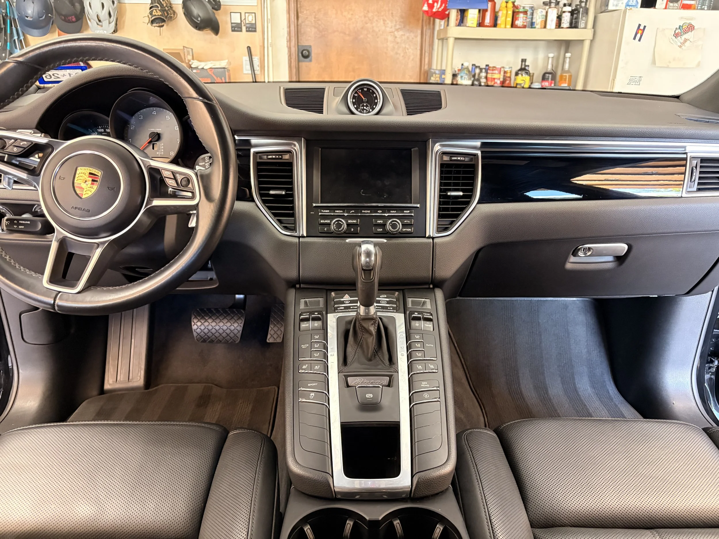Interior of a Porsche Macan car with a black dashboard, leather seats, and a steering wheel displaying the Porsche logo. View includes the instrument cluster, center console, and a clock on the dashboard.