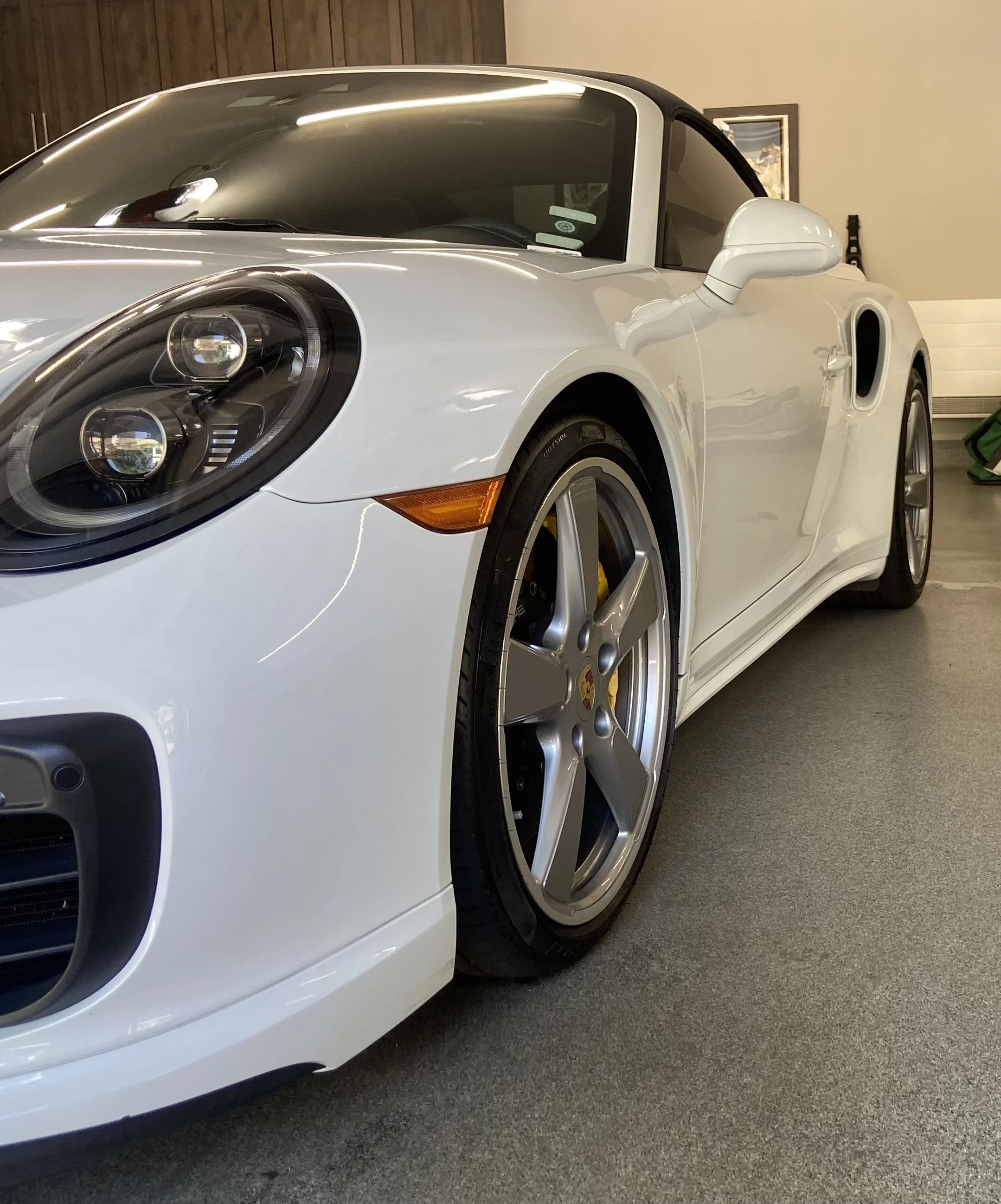 Close-up of a white sports car, focusing on the front left side, including the headlight, wheel, and side mirror, in an indoor setting.