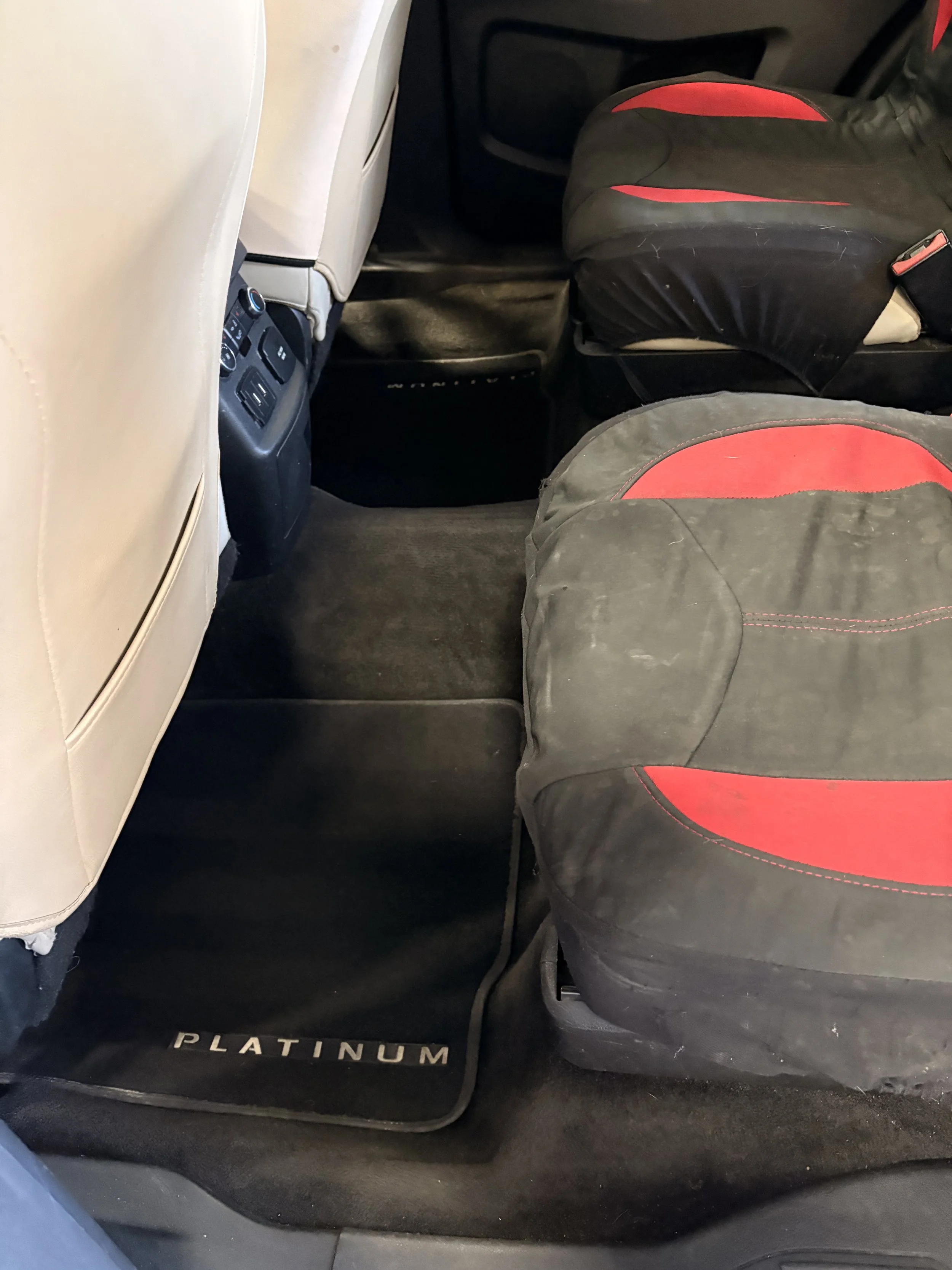 Inside a car showing beige seat, black floor mat with 'PLATINUM' embroidered, black and red car seat cover, and rear passenger area controls.