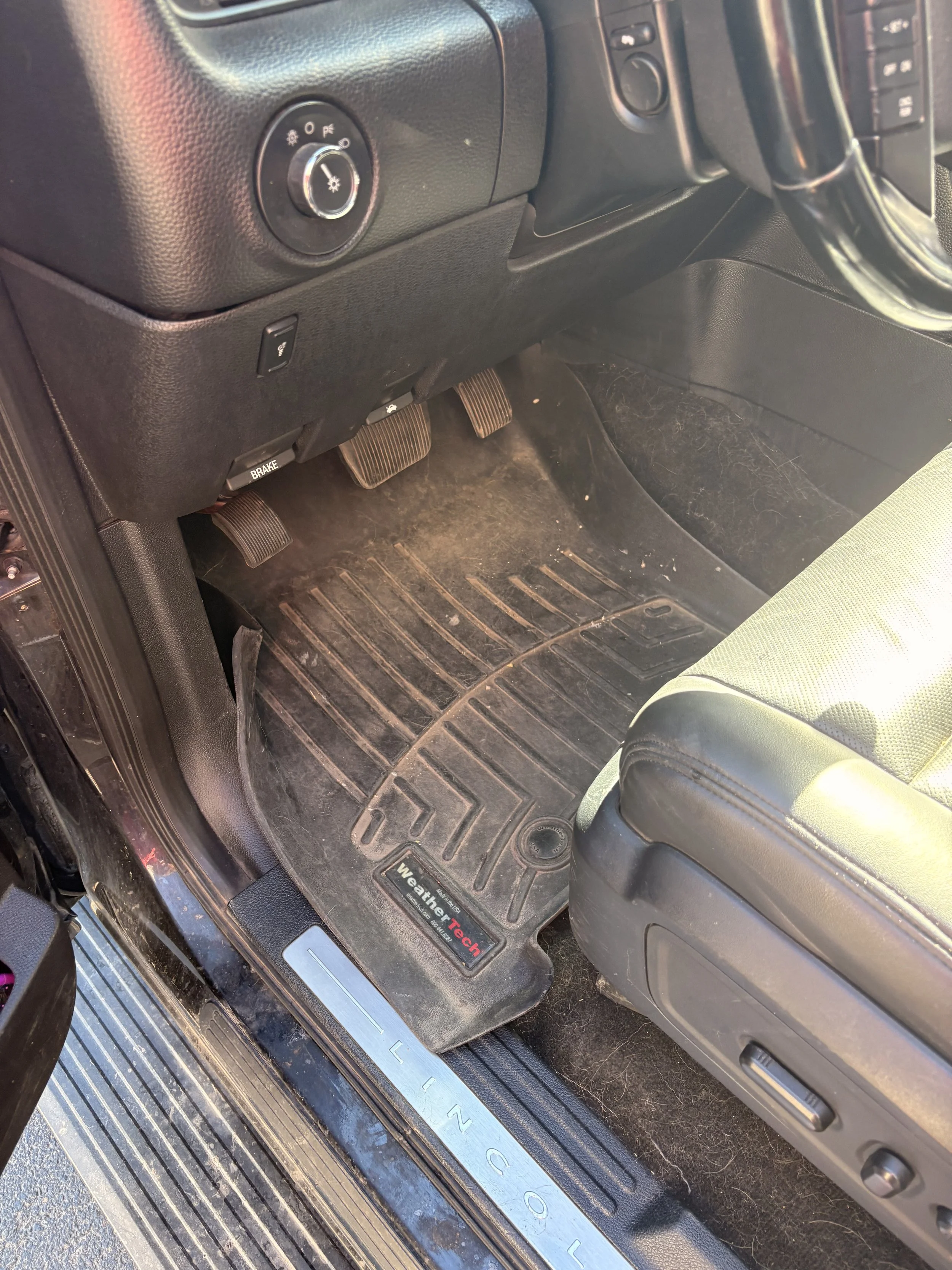 Interior of a vehicle showing the driver's side floor area with gas and brake pedals, a floor mat with a WeatherTech logo, and part of a green and black seat.