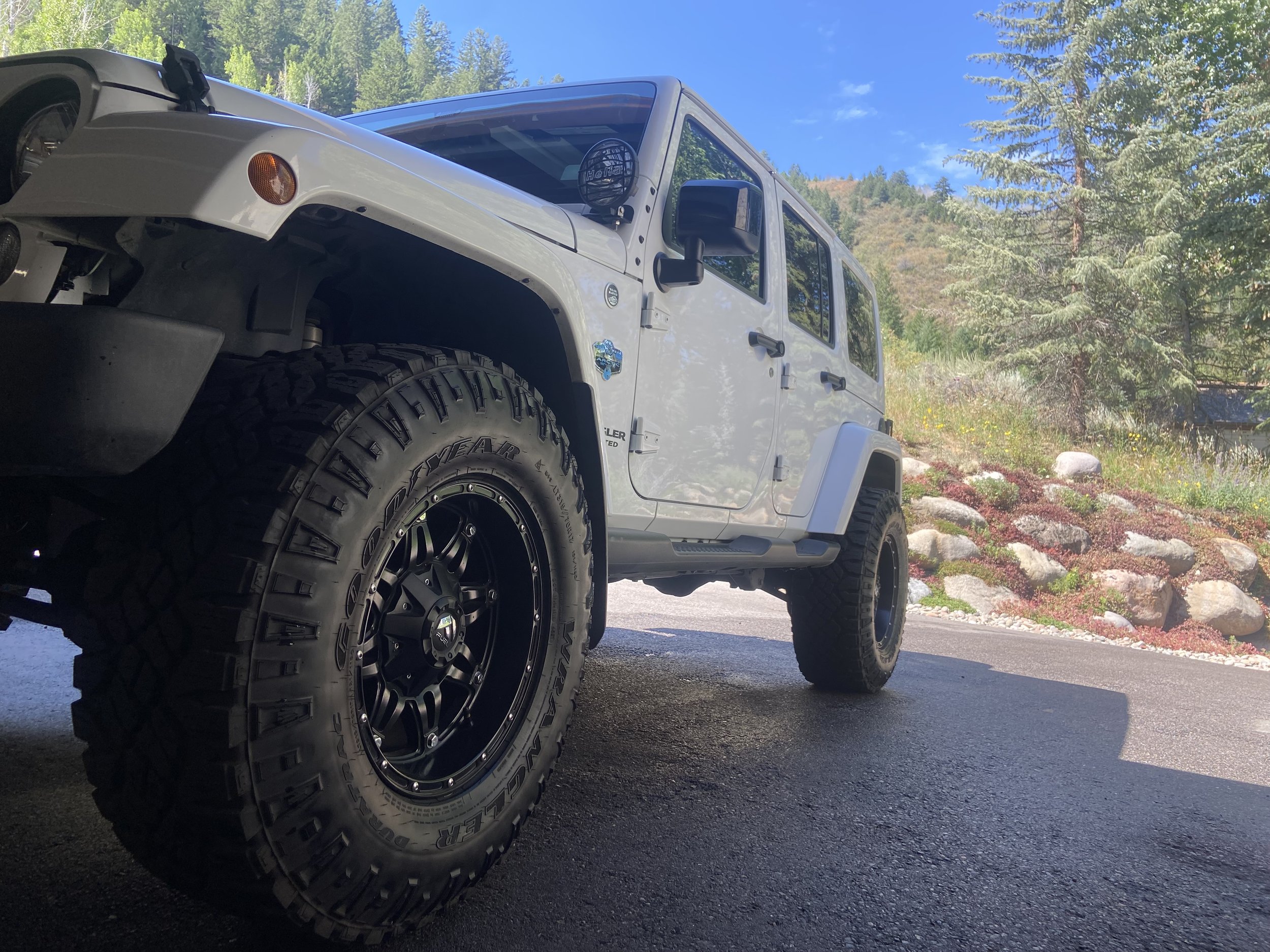 A white four-door Jeep parked on a paved lot surrounded by trees and rocks with a mountain in the background under a clear blue sky.