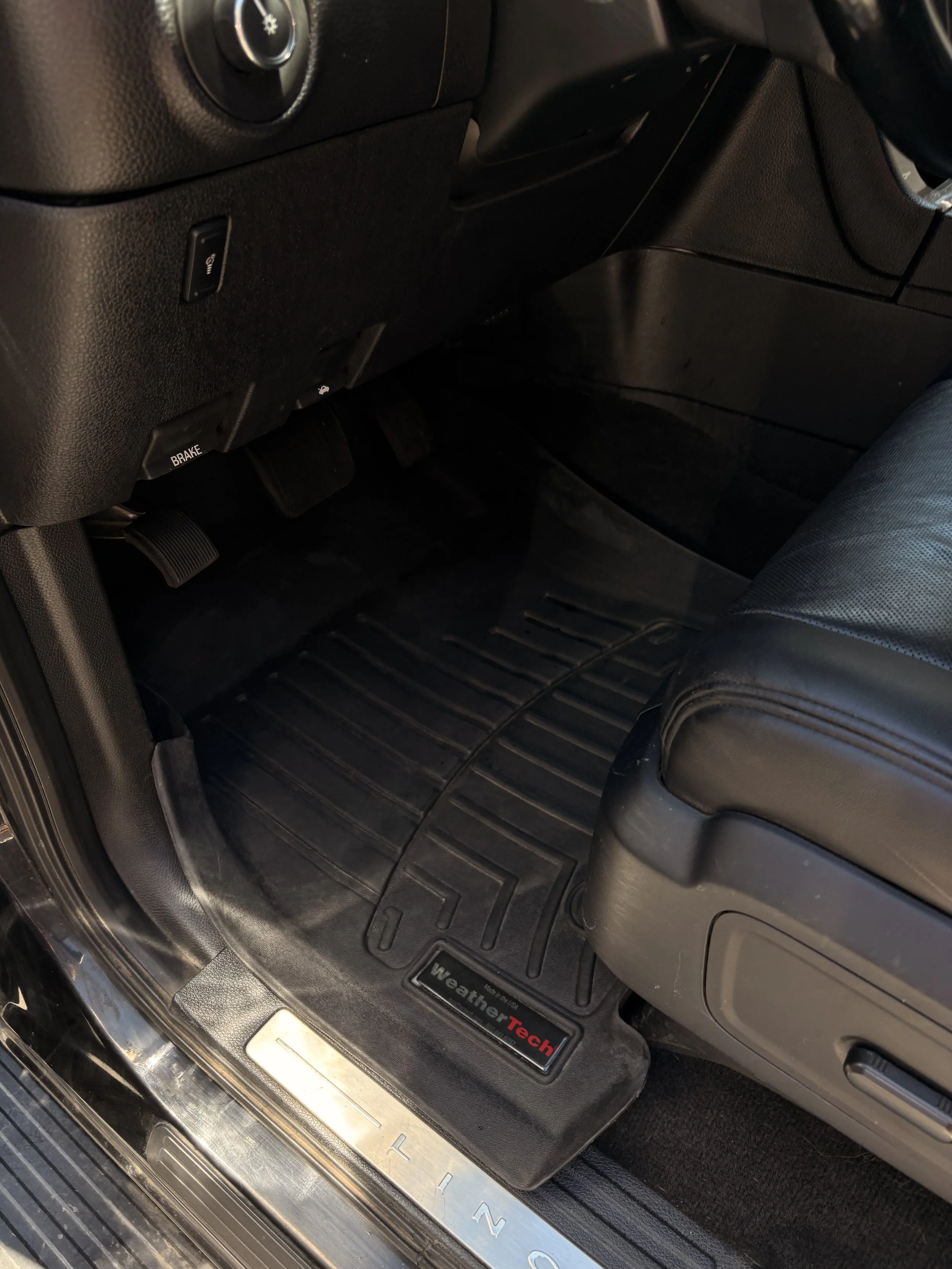 The interior of a vehicle showing the driver's side footwell with a black WeatherTech floor mat, a brake pedal, and part of the dashboard with a seat adjustment lever.