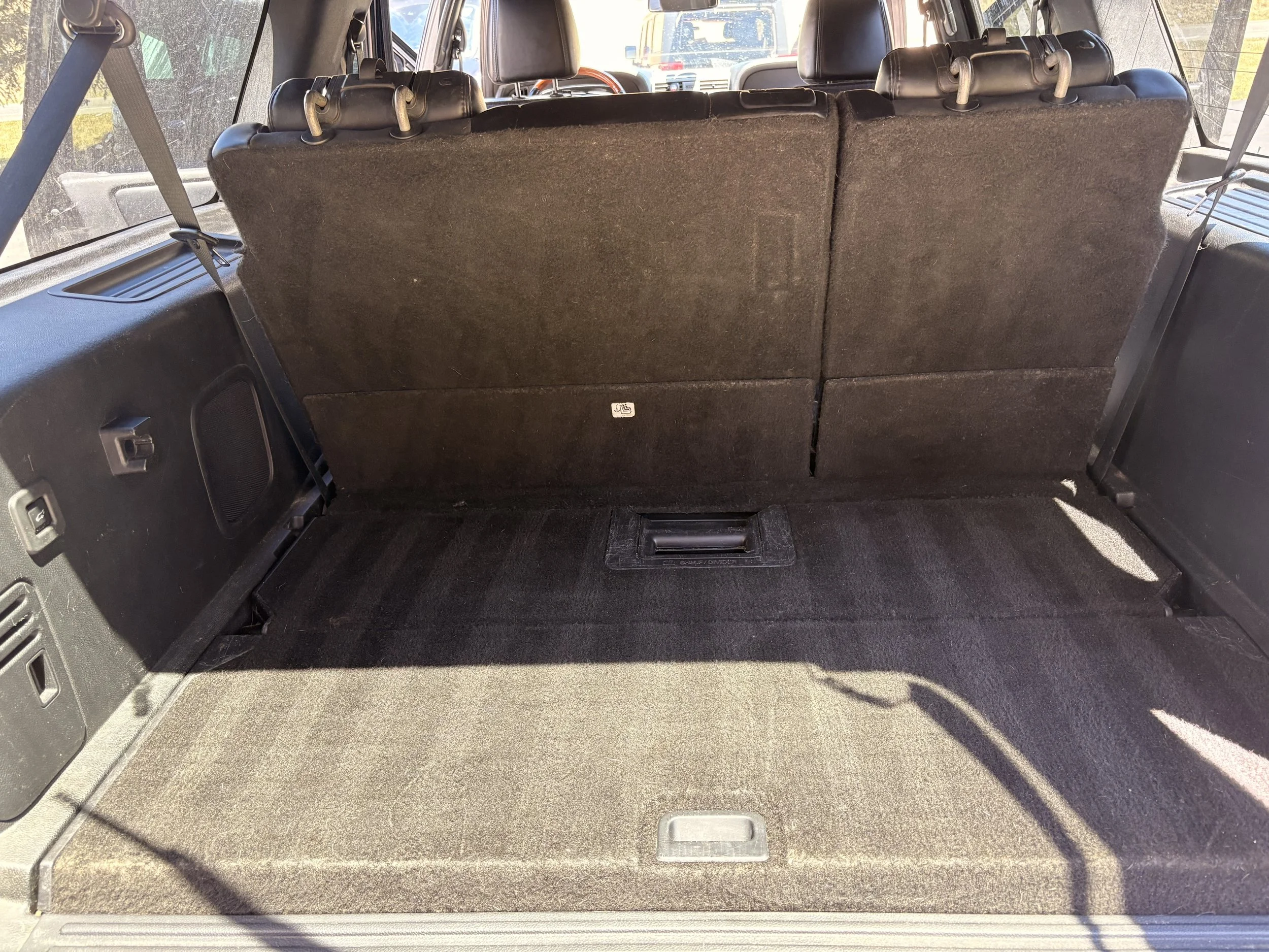 The image depicts the interior of a vehicle, showing the row of folded seats in the cargo area with a black carpeted floor and side panels. The seats are covered in dark fabric, and there are various electronic controls and vents visible along the si