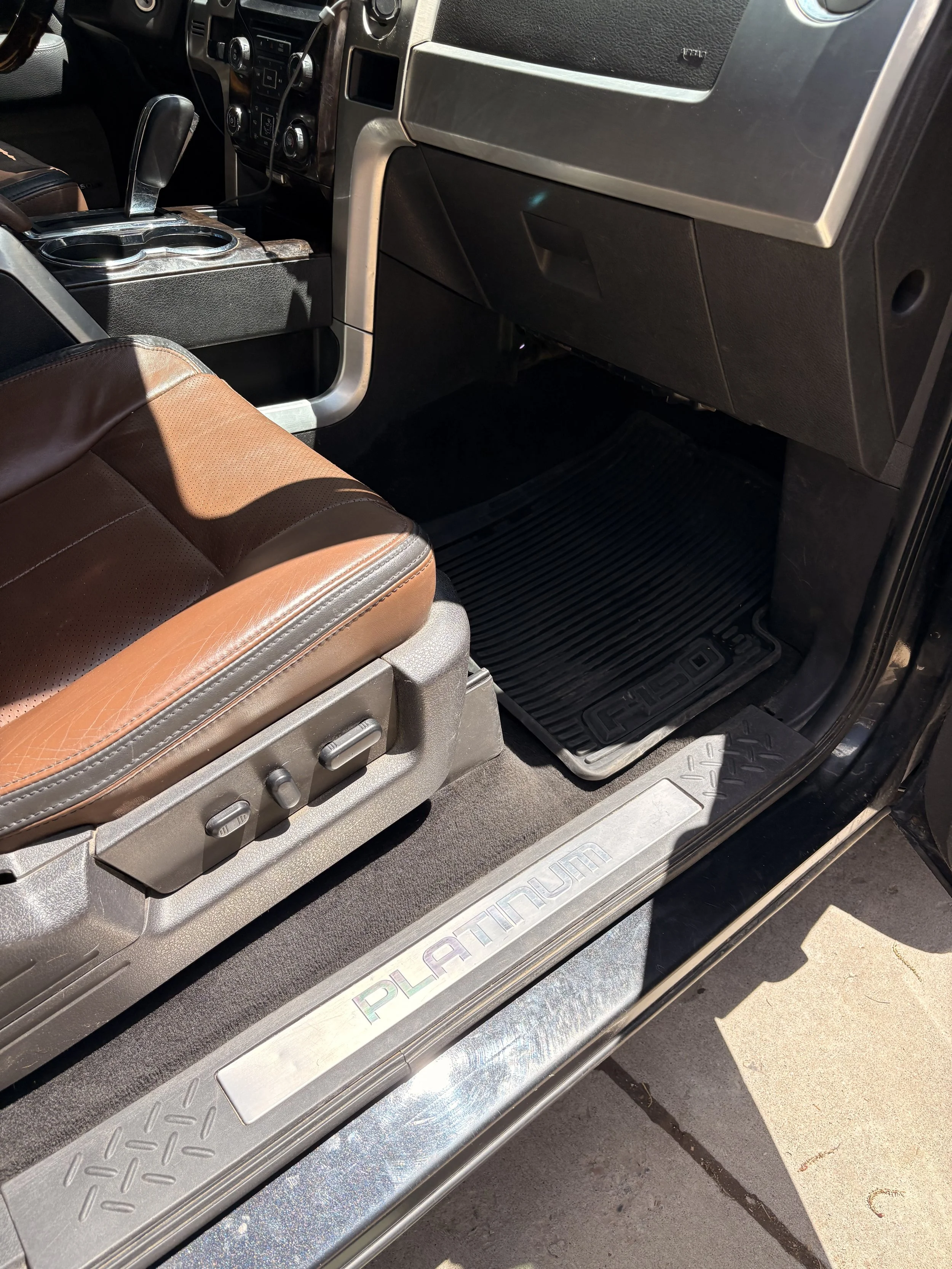 The interior of a vehicle showing the brown leather seat, gear shift, dashboard, and a black rubber floor mat with the word 'PLATINUM' on the door sill.