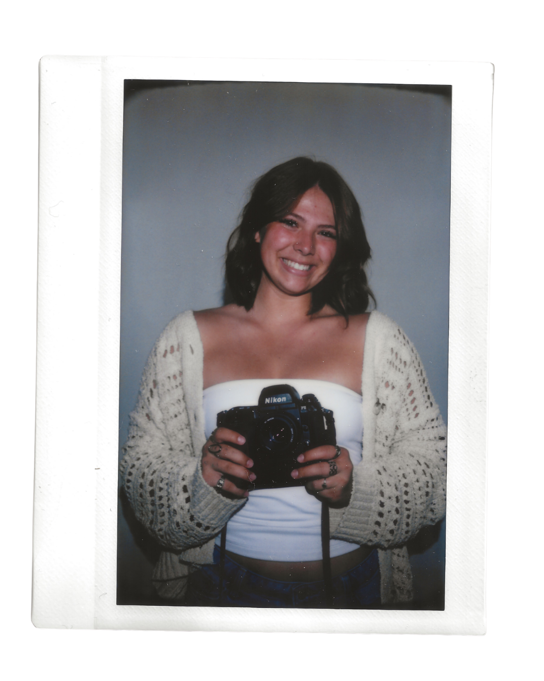 A Polaroid of a young woman with shoulder-length dark hair with a Nikon camera. She is smiling, wearing a white tube top, a beige open-knit cardigan, and several rings.