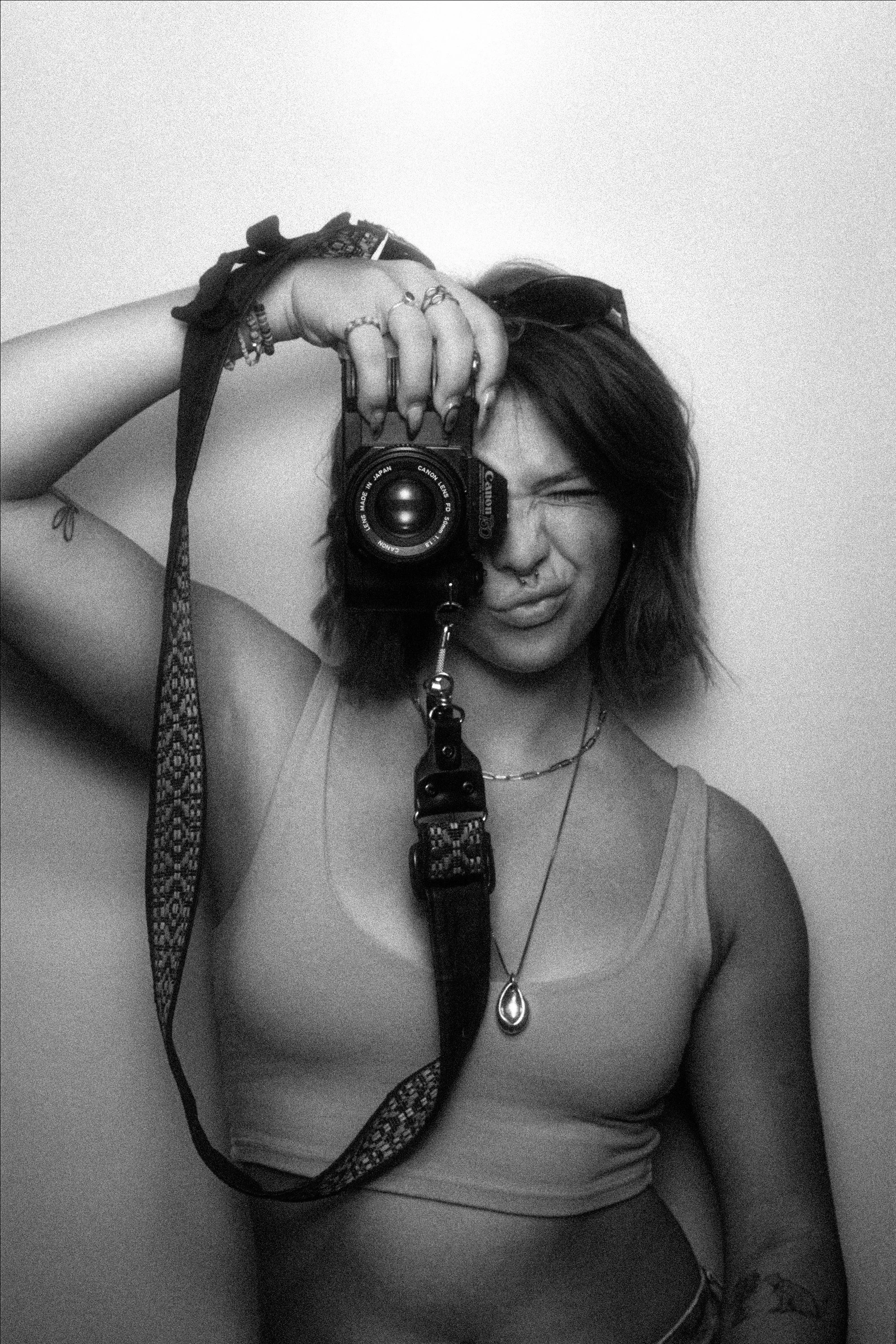 A woman with wavy hair taking a photo of herself in a mirror with a camera. She is wearing a tank top and jewelry, with a curtain or fabric seen on her right arm, and a plain wall as background.