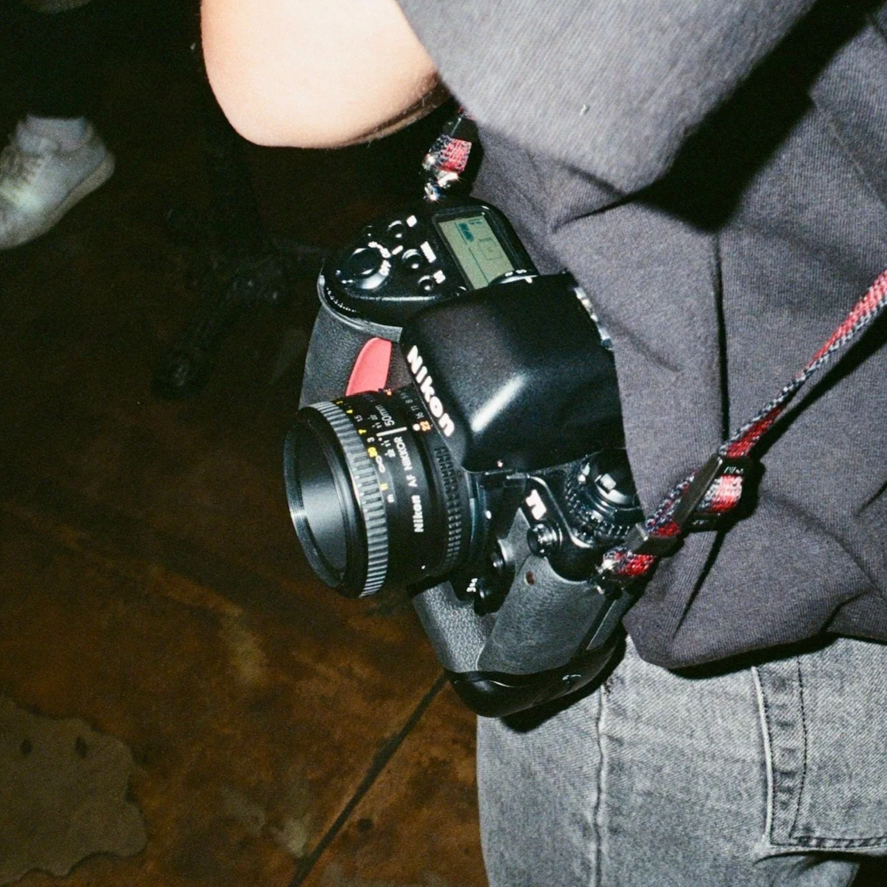 A Nikon DSLR camera with a 50mm lens resting on a person's lap, who is wearing gray jeans and a gray top. The camera strap is red with black accents.