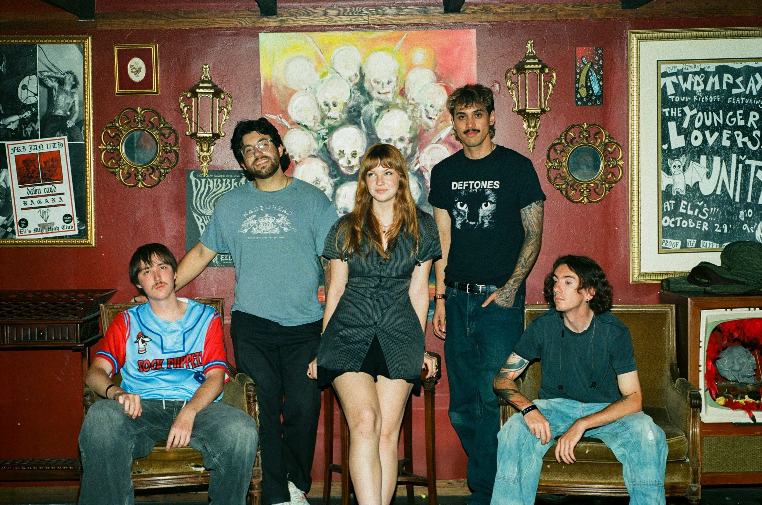 A group of five young people posing in an art-filled room with red walls. Four are standing behind a woman with red hair in the center, seated in front. The background features paintings, mirrors, and posters.
