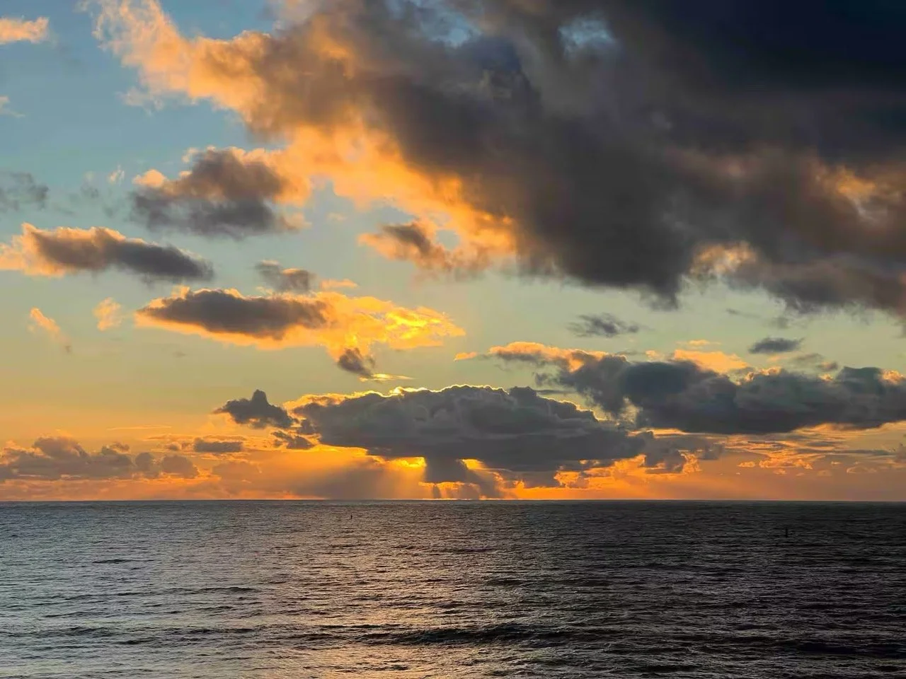Sunset over the ocean with scattered dark clouds and a golden glow near the horizon.
