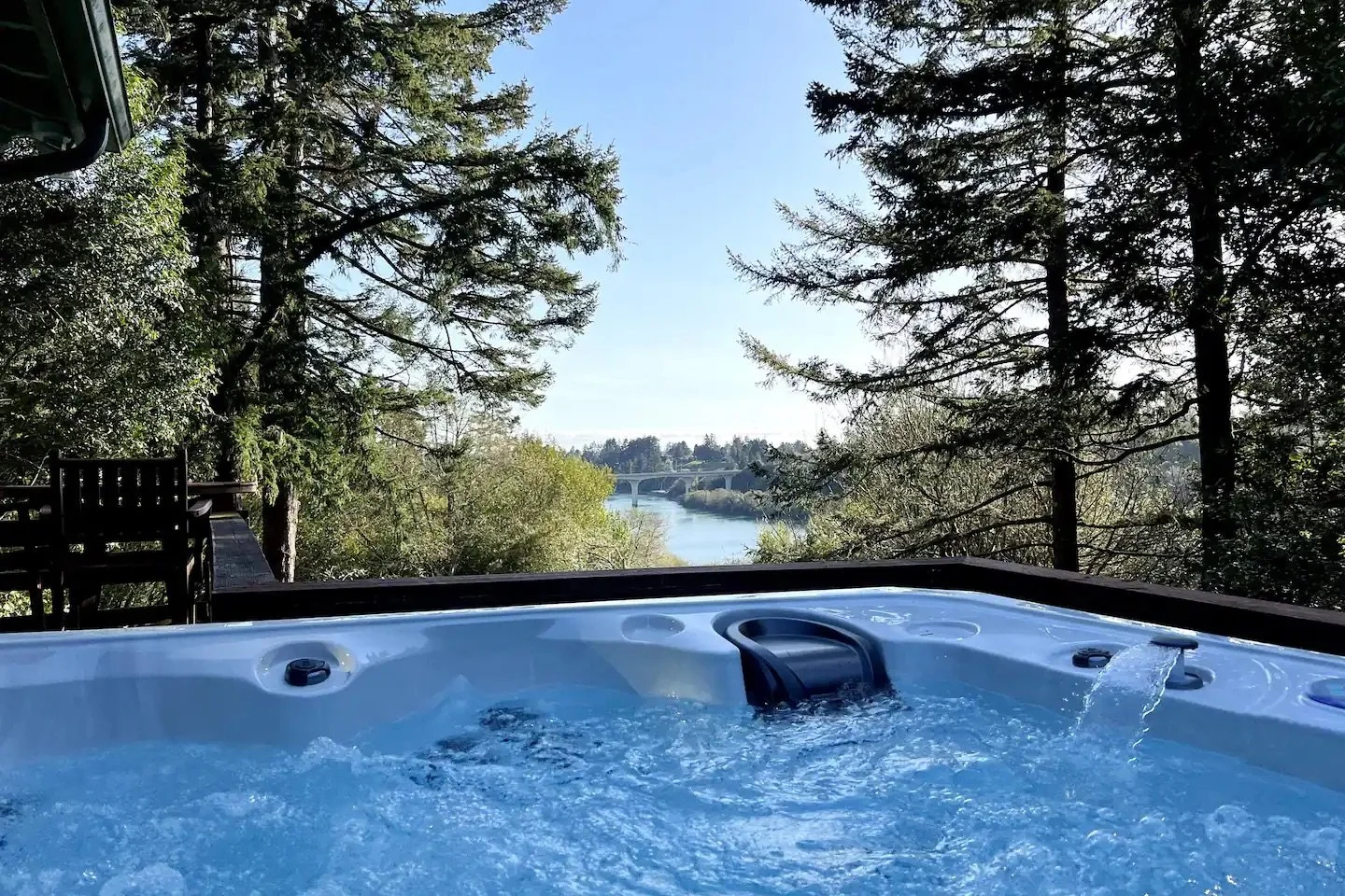 A hot tub on a deck overlooking a river and a bridge, surrounded by trees, on a clear day.