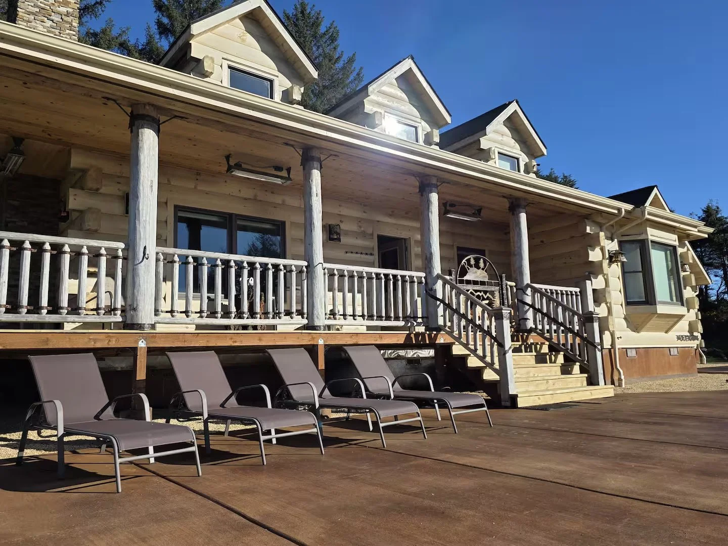 A wooden house with a porch featuring six large wooden columns and a stairway leading up to it. There are four lounge chairs on the deck in front of the house, and the house has multiple windows and dormers on the roof. The sky is clear and blue.
