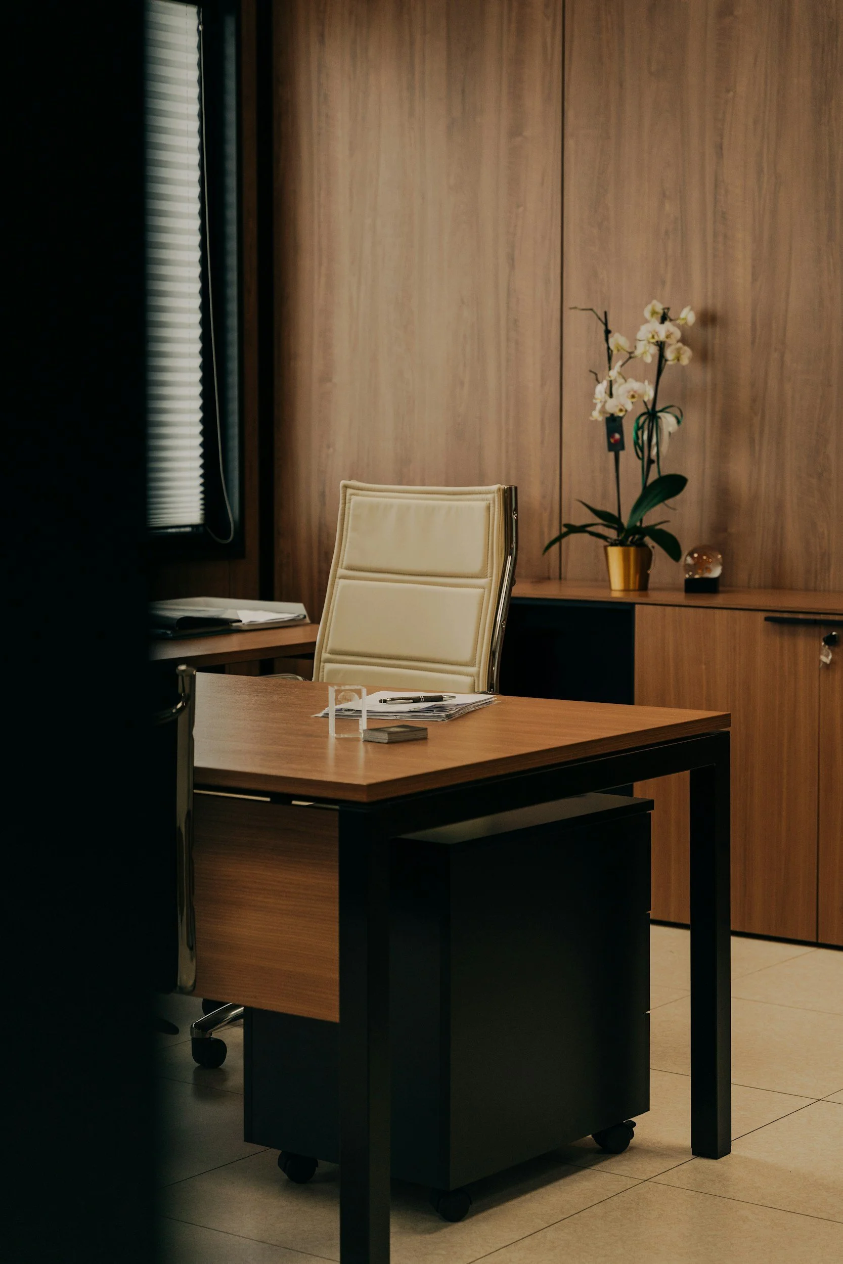 An office with a wooden desk and cream-colored office chair. On the desk, there are some papers, a pen, and a tablet. Behind the desk, there is a wooden counter with a plant in a yellow pot, a small decorative object, and a lamp. To the left, there is a window with blinds partially open, allowing some light into the room.