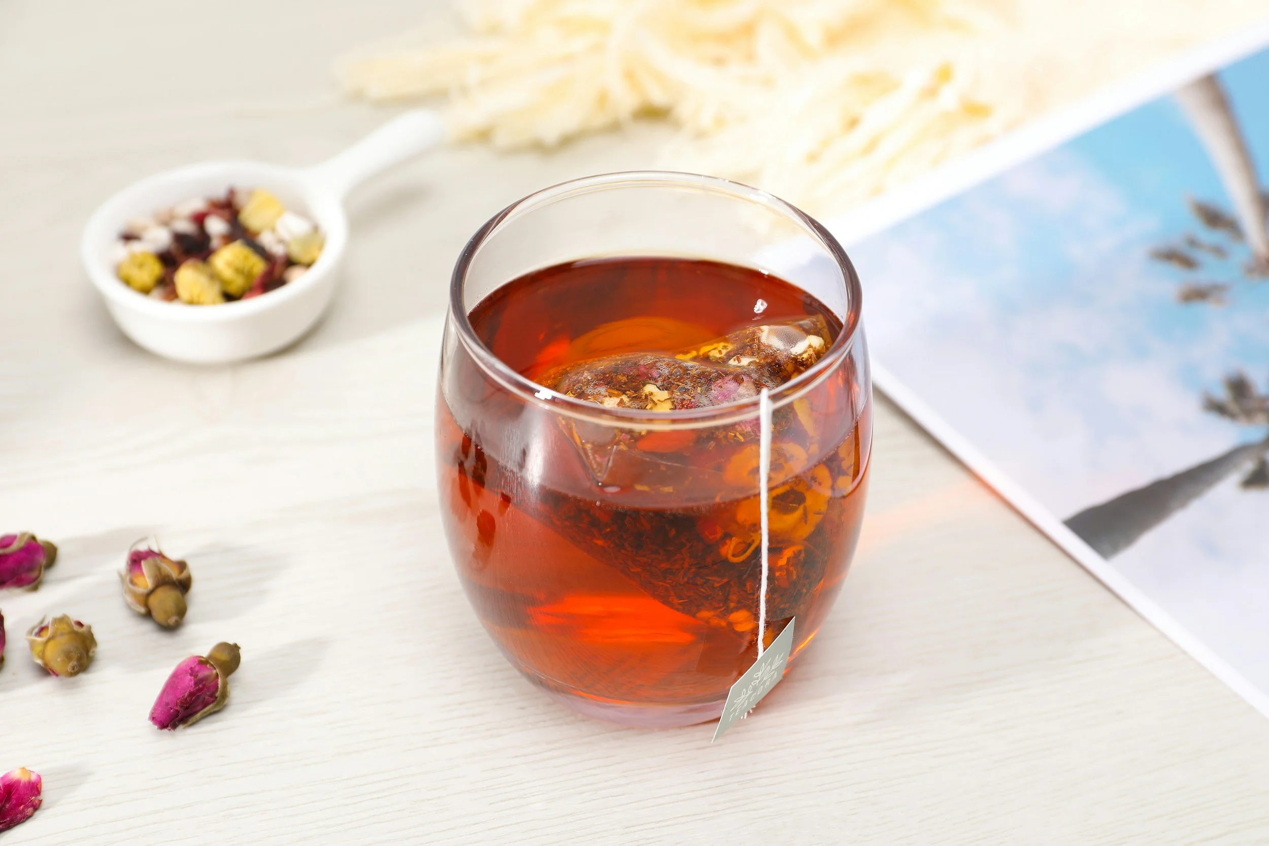 A glass of iced tea with a tea bag inside, placed on a white surface. In the background, there is a bowl with dried flowers or herbs and some scattered pink rosebuds.