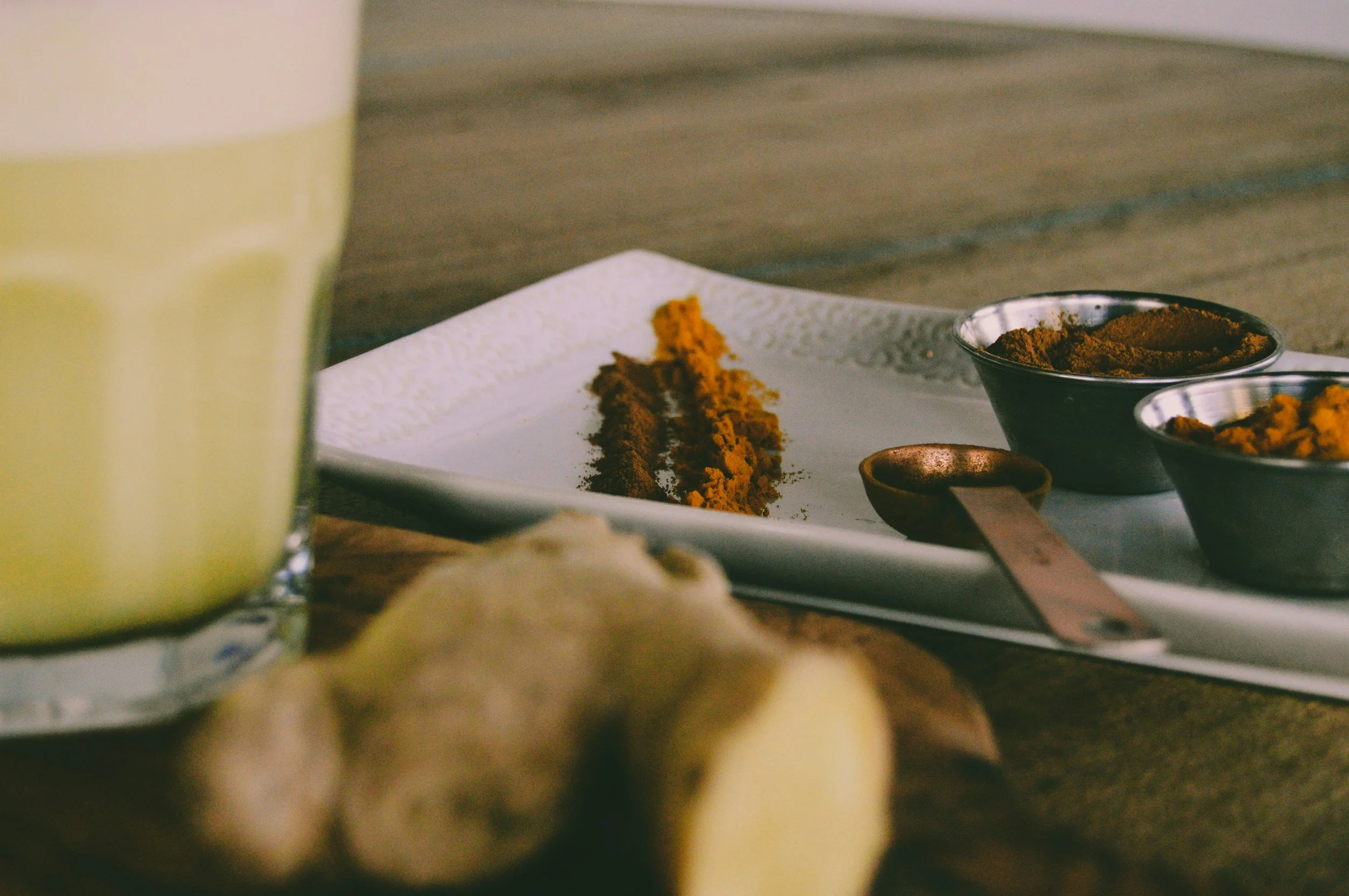 Close-up of a white rectangular plate with a smear of bright orange spice or paste, surrounded by small black and silver bowls containing different spices, on a wooden surface. The foreground shows a blurred glass of yellow beverage and some chopped ginger.