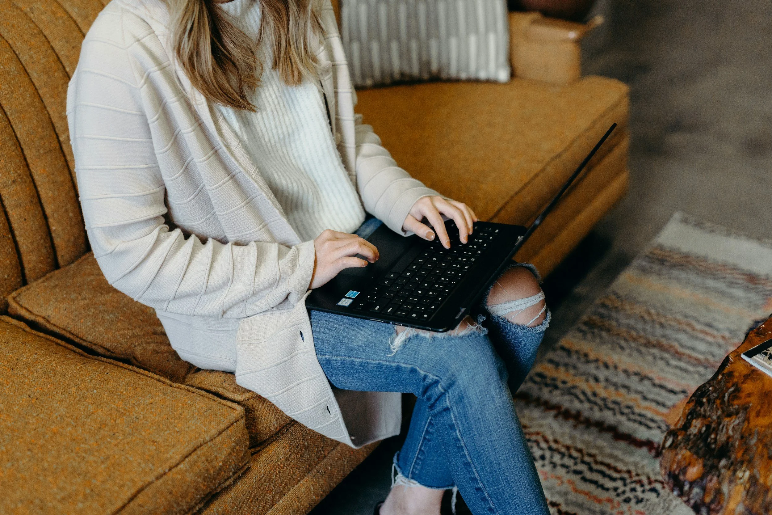 A person with long hair, wearing a white striped sweater and ripped jeans, sitting on a brown upholstered bench, using a black laptop.