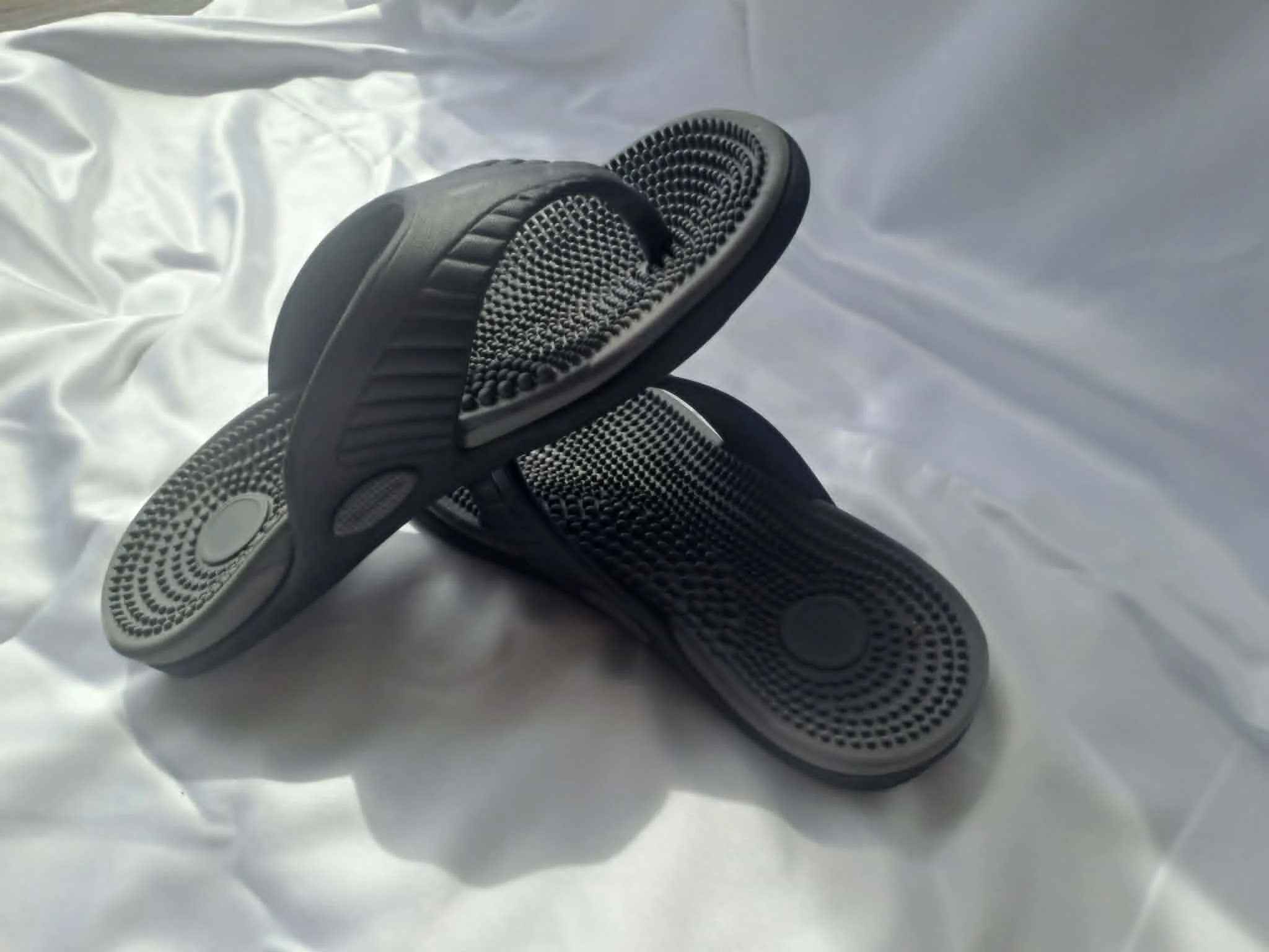 Two black textured slide sandals placed on a white crumpled fabric surface.