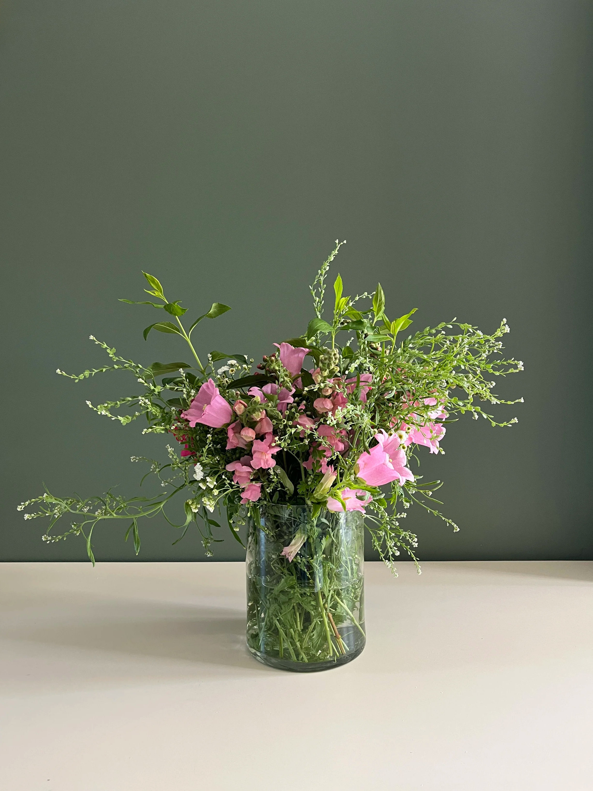 A tall glass vase with a bouquet of pink, white, and green flowers and greenery, placed on a white surface against a dark gray background.