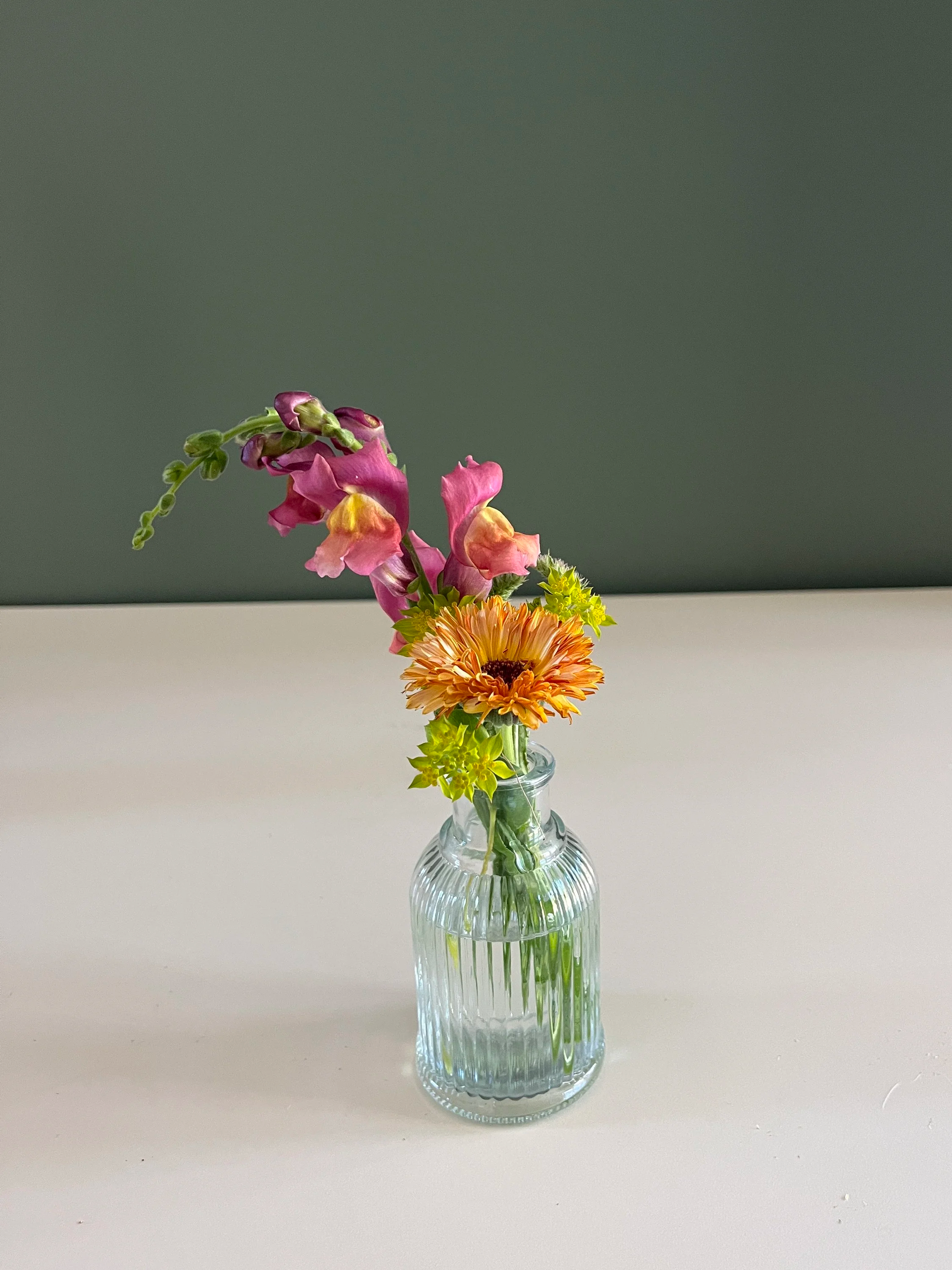 A small bouquet of colorful flowers in a clear, ribbed glass vase on a white surface with a gray wall background.