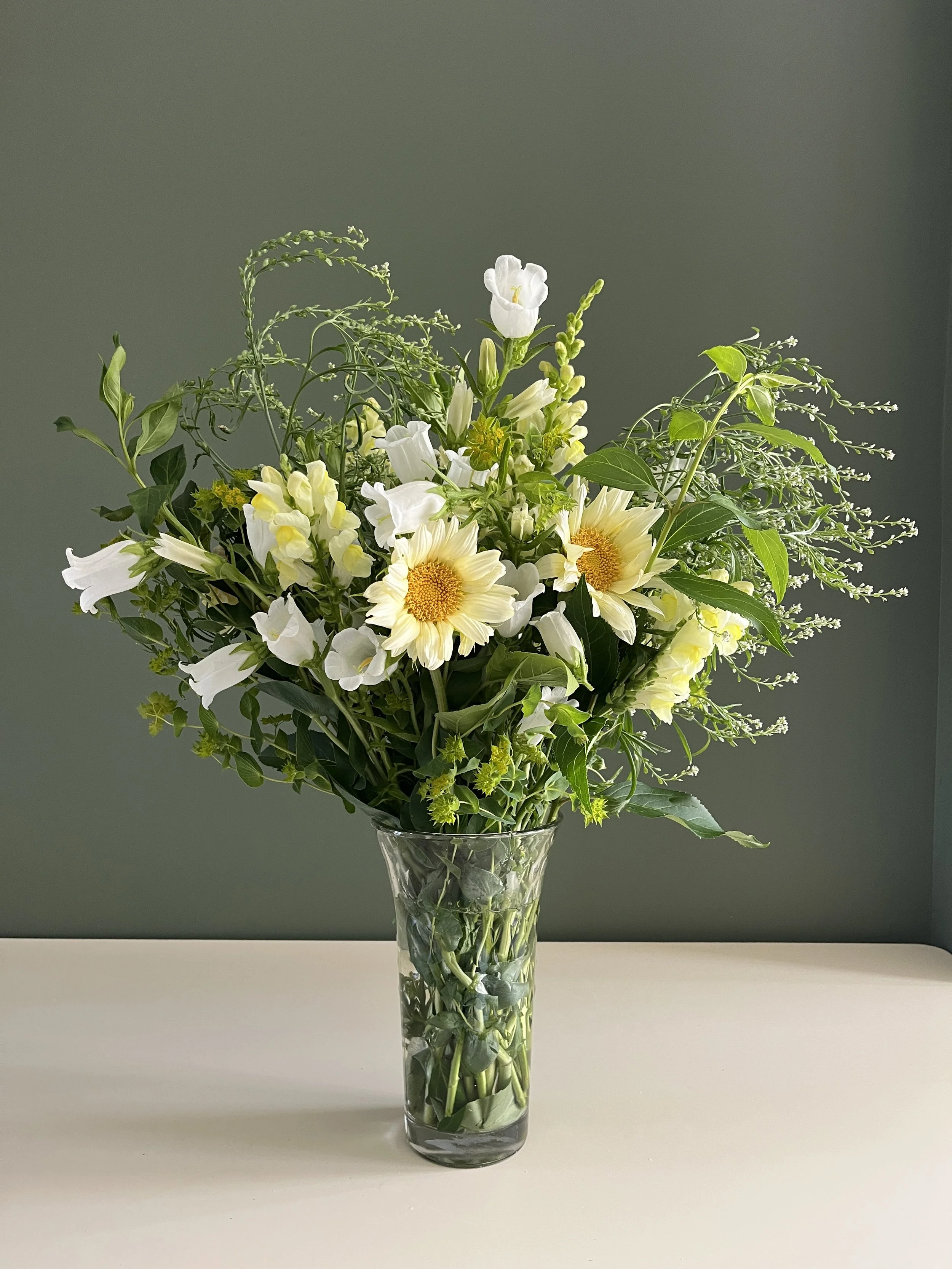 A bouquet of white and yellow flowers in a tall, clear glass vase on a white surface, against a plain gray background.
