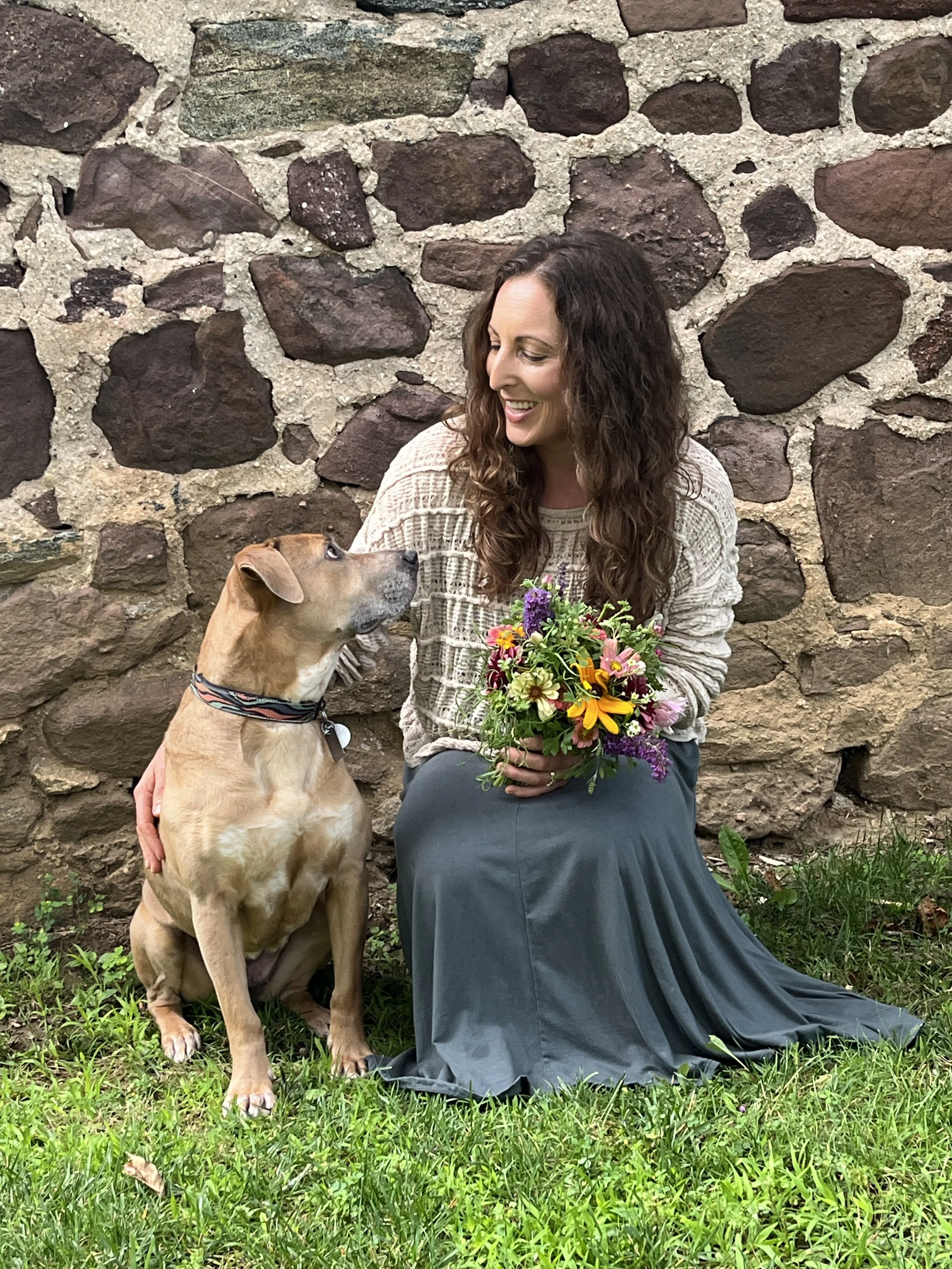 A woman with long curly brown hair holding a colorful bouquet of flowers while smiling at a large tan dog with a black collar sitting beside her against a stone wall background.