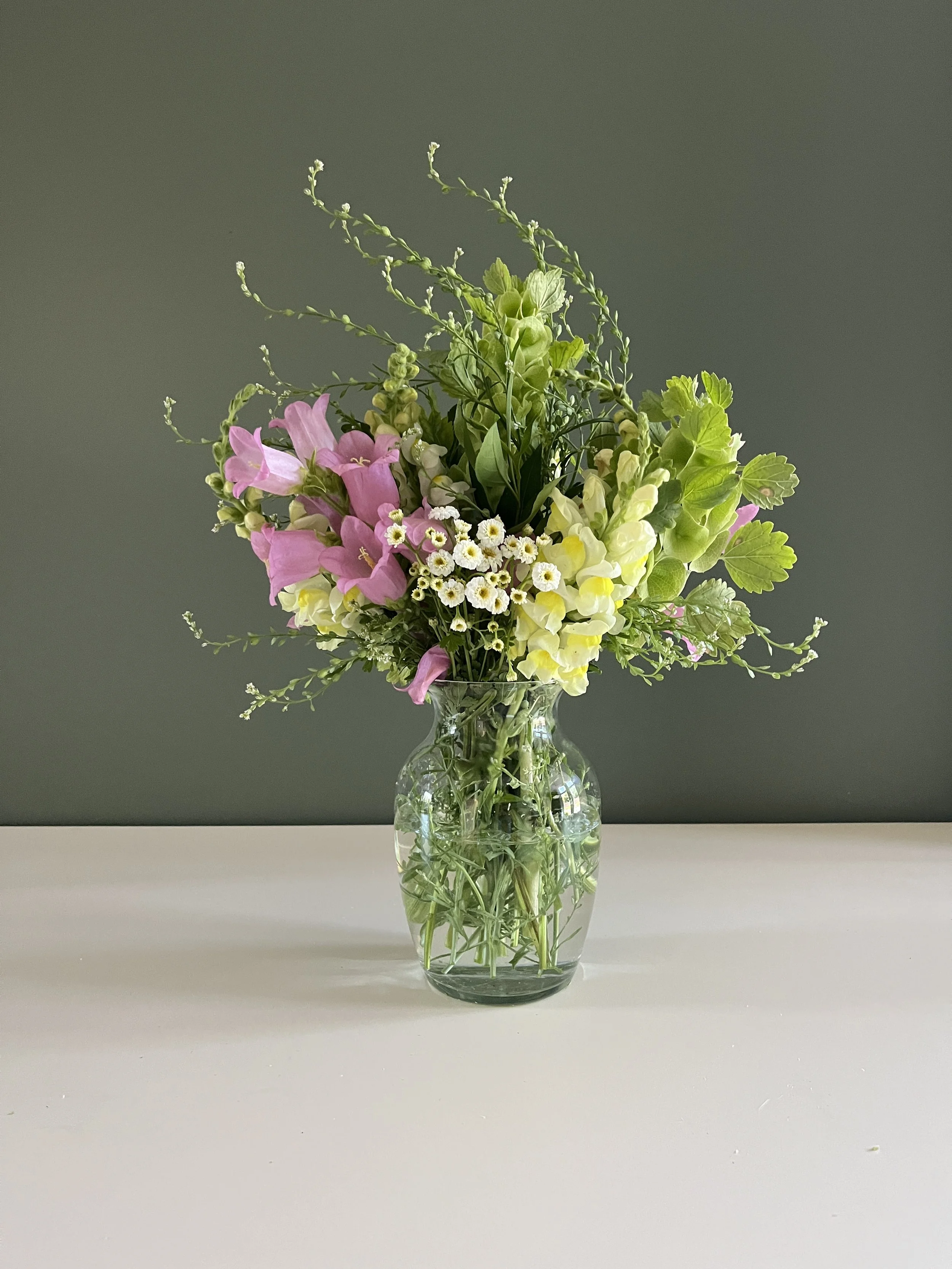 A glass vase with a mixed bouquet of pink, white, and yellow flowers, along with green leaves and stems, on a white surface against a neutral gray background.