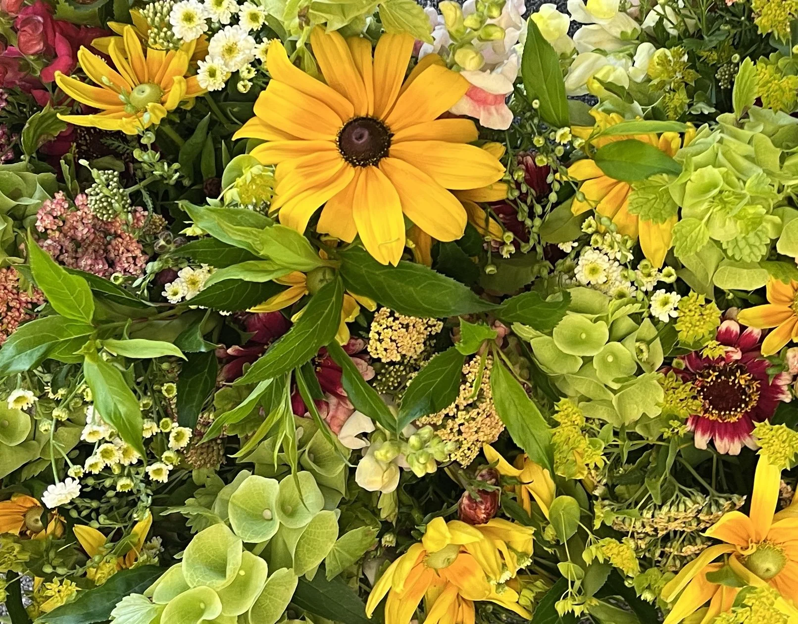 Close-up of a vibrant assortment of various flowers and green leaves, including yellow daisies, white small blossoms, pink flowers, and green hydrangea blooms.