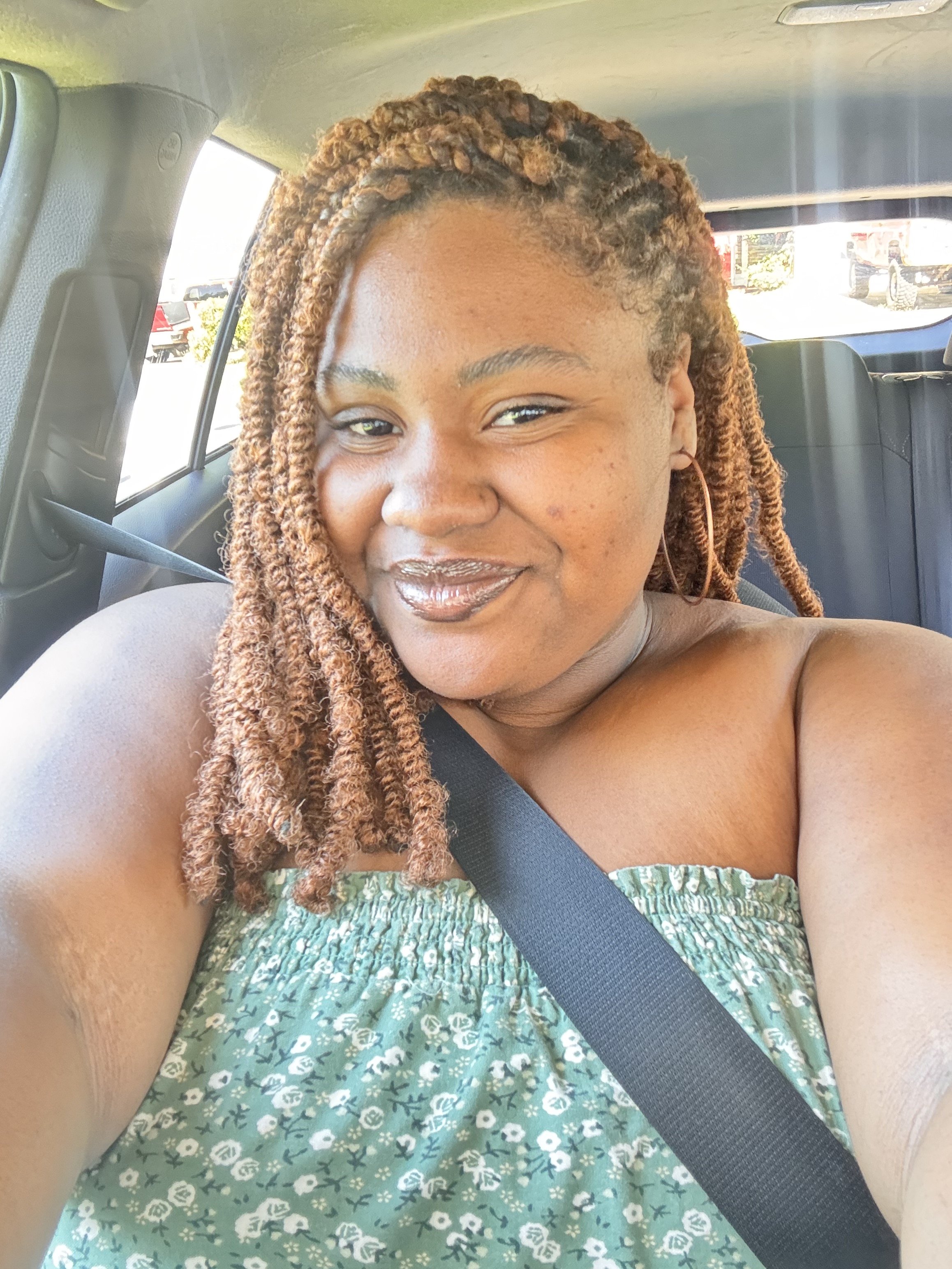 A woman with brown, curled hair sitting in a car, smiling, wearing a green patterned strapless top, with a seatbelt across her chest.