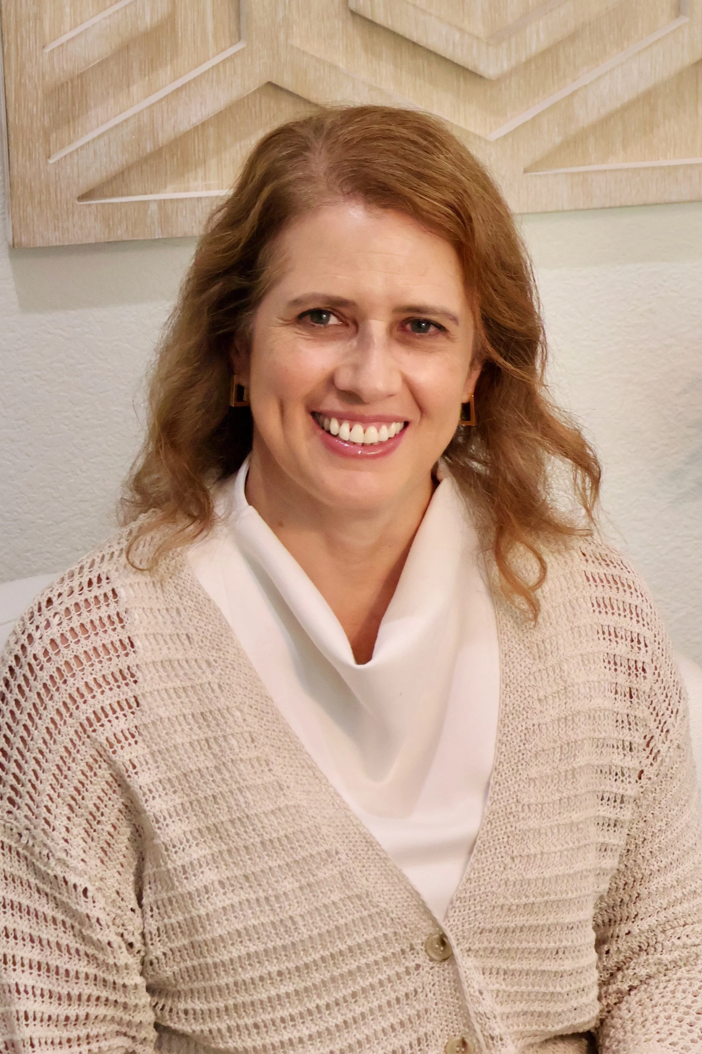 A smiling woman with brown hair, wearing earrings, a white collared shirt, and a beige, open-knit cardigan, sitting in front of a textured wall with a wooden art piece.