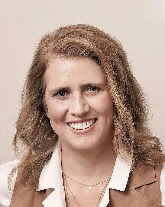 A smiling woman with shoulder-length wavy blonde hair wearing a light-colored shirt and brown blazer, standing against a plain background.